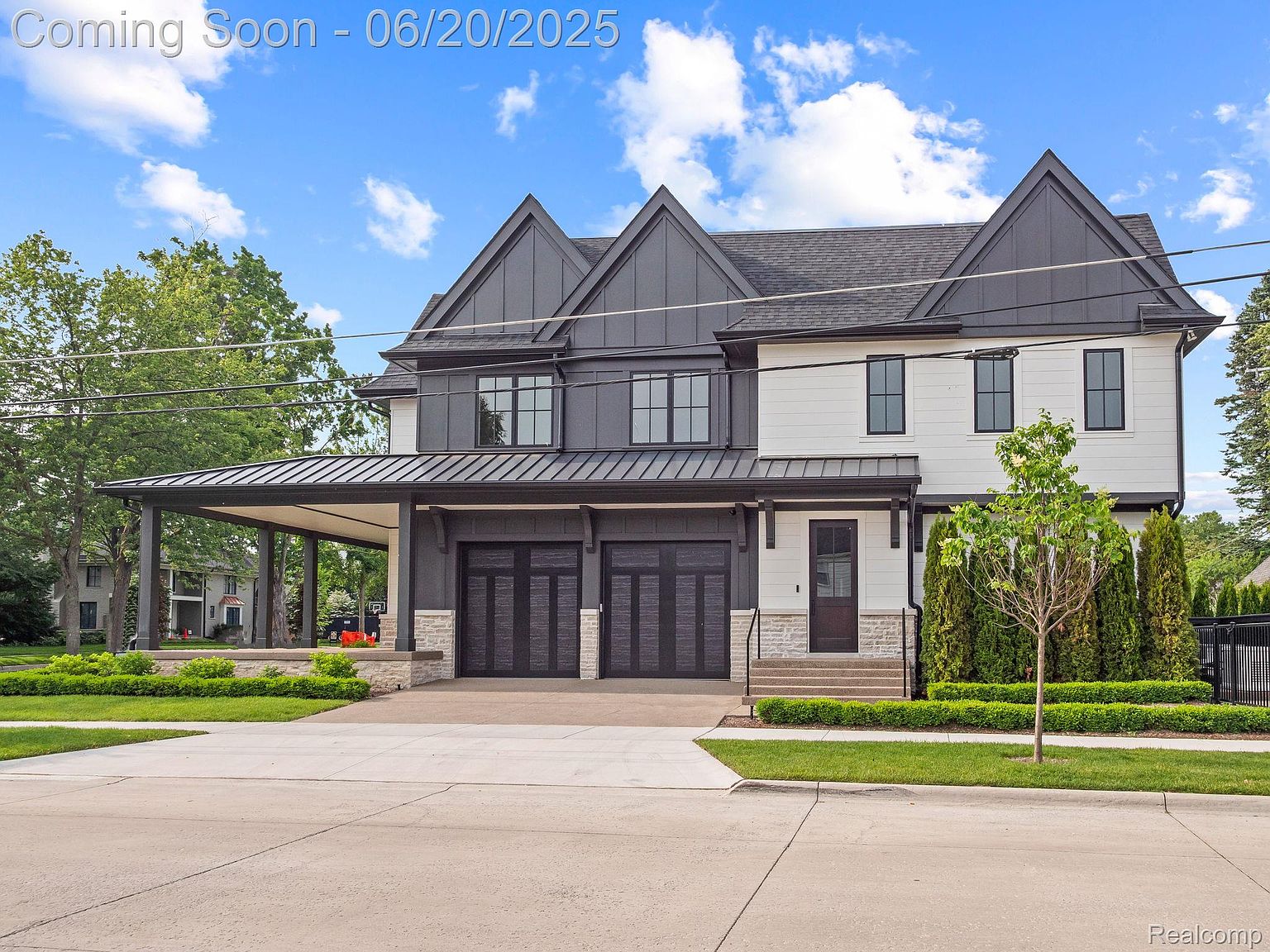 This is a front exterior view of a modern two-story home. The house features a combination of dark gray and white siding, with black framed windows and a dark gray roof. A covered carport extends from the side of the house, leading to a two-car garage with dark wooden doors accented by stone pillars. The landscaping is well-maintained with green lawns, trimmed hedges, and young trees, enhancing the property's curb appeal.