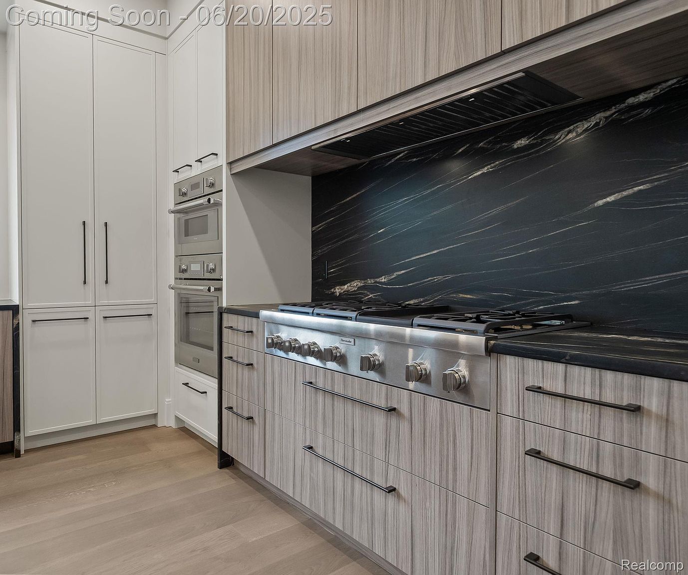This is a modern kitchen featuring sleek, light wood-grain cabinetry with long, dark hardware. A stainless steel range with a black marble backsplash is the focal point, complemented by built-in ovens and white cabinets on the left. The kitchen exudes a sophisticated and contemporary aesthetic.