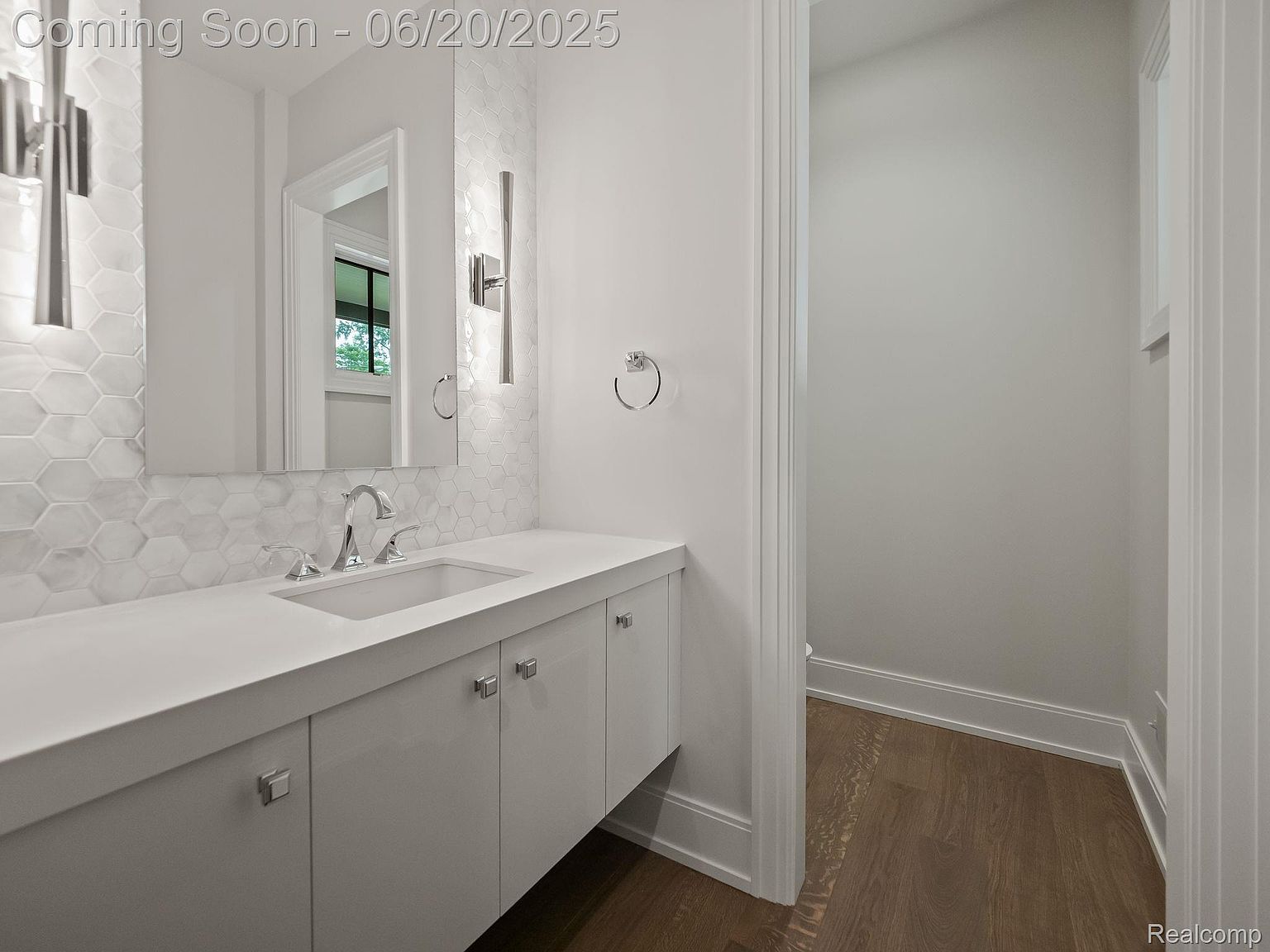 This is a well-lit guest bathroom featuring a modern vanity with white cabinetry and a white countertop. A stylish mirror hangs above the sink, complemented by unique sconces and hexagonal tile backsplash. The room also includes a doorway leading to another space, showcasing hardwood flooring.