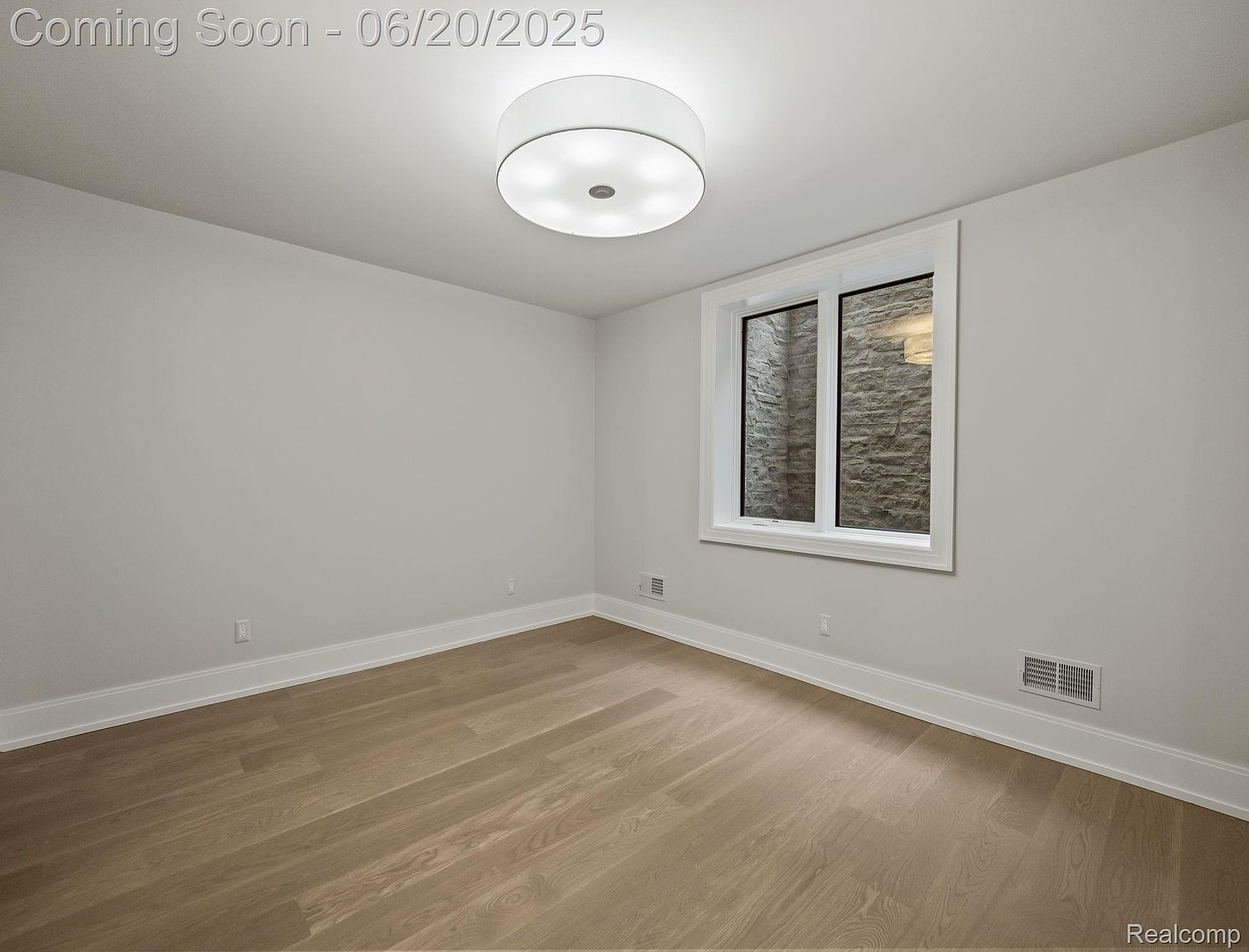 The image showcases a well-lit, modern bedroom with light hardwood flooring and neutral-colored walls. A window provides natural light and a view of a stone wall outside. The room features a contemporary ceiling light fixture and minimalist design, creating a clean and inviting space.