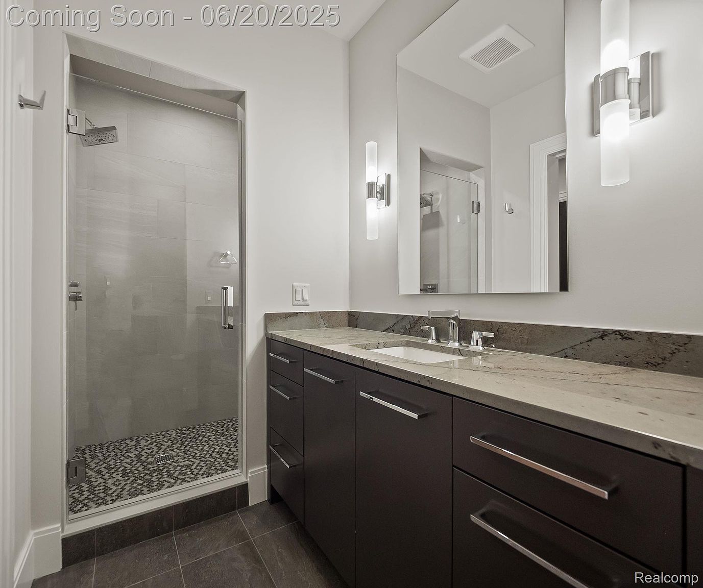 This is a primary bathroom featuring a walk-in shower with glass doors and a mosaic tile floor. The vanity has dark wood cabinets, a light-colored countertop with dark veining, and modern hardware. A large mirror reflects the shower and a sconce, enhancing the sense of space and light.