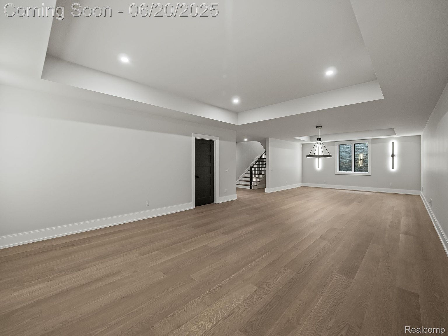This is a spacious living room featuring light hardwood floors and neutral gray walls. The room has recessed lighting and a modern chandelier, with a staircase visible in the background. The overall impression is clean, bright, and modern, suggesting a newly constructed or recently renovated space.