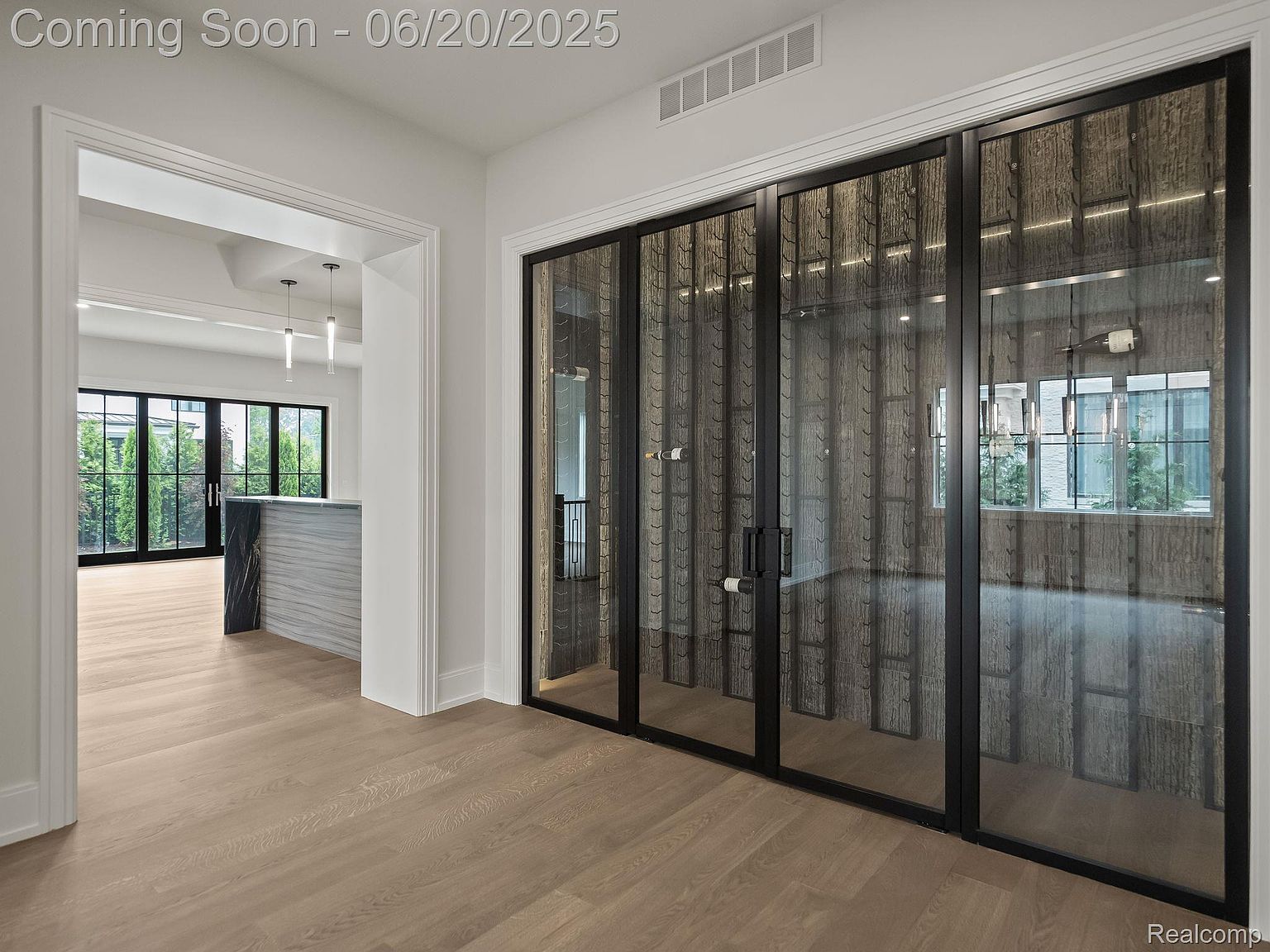 This interior shot showcases a modern wine cellar with a glass-enclosed design and black framing. The cellar features unique wine storage racks and is well-lit, creating an elegant and sophisticated space. The light wood flooring adds warmth to the room, and the view through the doorway suggests an open floor plan.