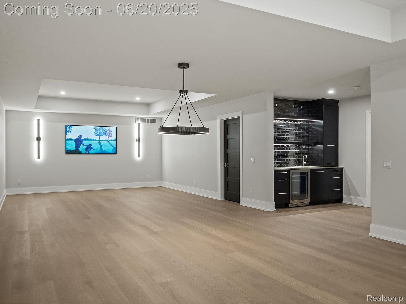 This is an interior shot of a modern living room featuring light hardwood floors and neutral gray walls. A built-in bar area with dark cabinetry and a tiled backsplash is visible, along with a large television mounted on the wall. The room is illuminated by recessed lighting and a contemporary chandelier, creating a bright and inviting atmosphere.