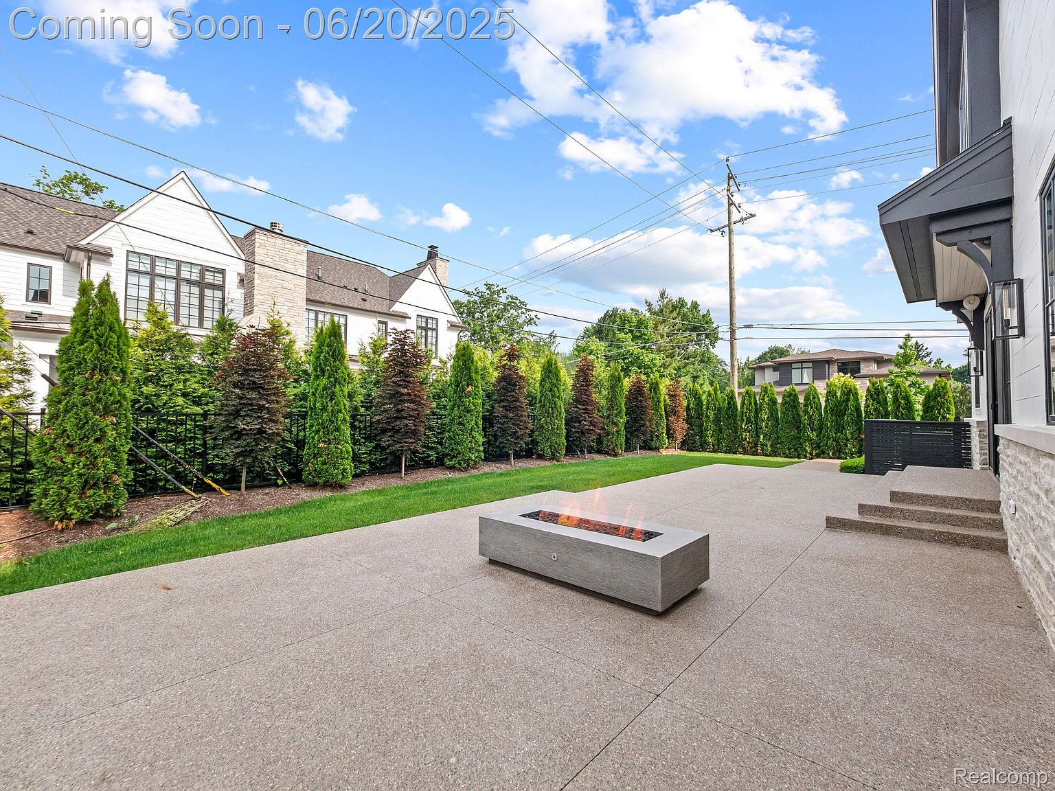This image showcases a modern patio area with a sleek, rectangular fire pit as a focal point. The patio is constructed from a light-colored concrete material, and is surrounded by lush green landscaping, including tall, manicured trees and a well-maintained lawn. The backdrop features a stylish home with a combination of white siding and stone accents, creating an inviting outdoor living space.
