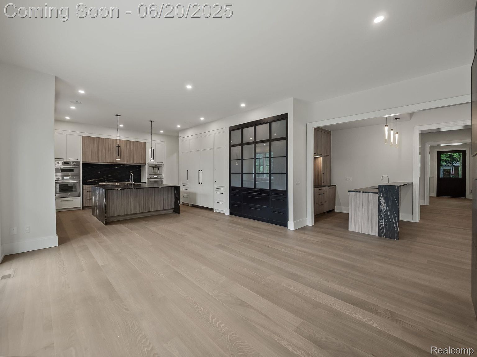 This is a spacious, modern kitchen featuring a large island with a dark countertop and light wood cabinetry. The kitchen includes stainless steel appliances, white cabinetry, and a black backsplash. The open floor plan leads to other areas of the house, creating a seamless flow.
