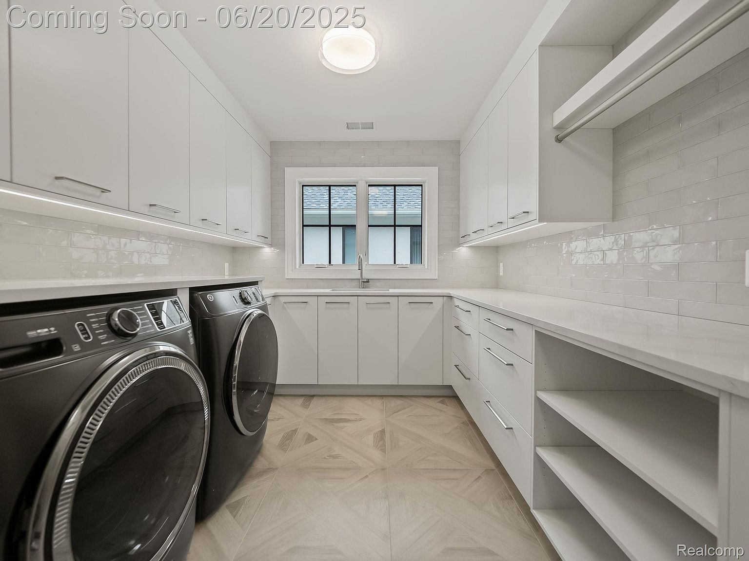 This is a modern laundry room featuring sleek, white cabinetry and countertops, complemented by light-colored herringbone-patterned flooring. A front-loading washer and dryer set are prominently displayed, and a window provides natural light. The overall design is clean, bright, and functional, creating an appealing space for laundry tasks.