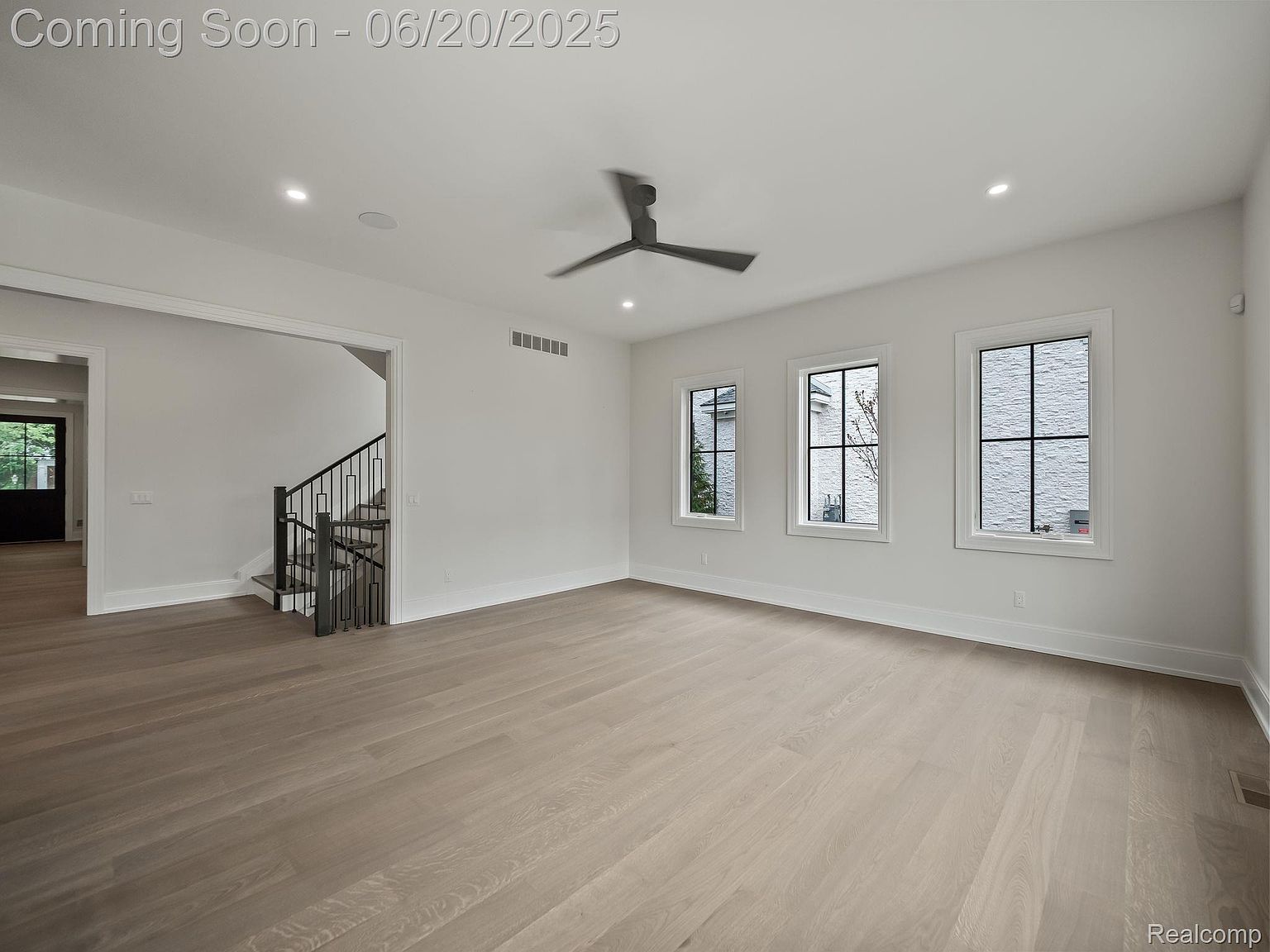 This is an interior shot of a spacious living room featuring light hardwood flooring and white walls. Three windows provide ample natural light, and a modern ceiling fan adds a touch of contemporary style. A staircase is visible through an adjacent doorway, suggesting an open floor plan.
