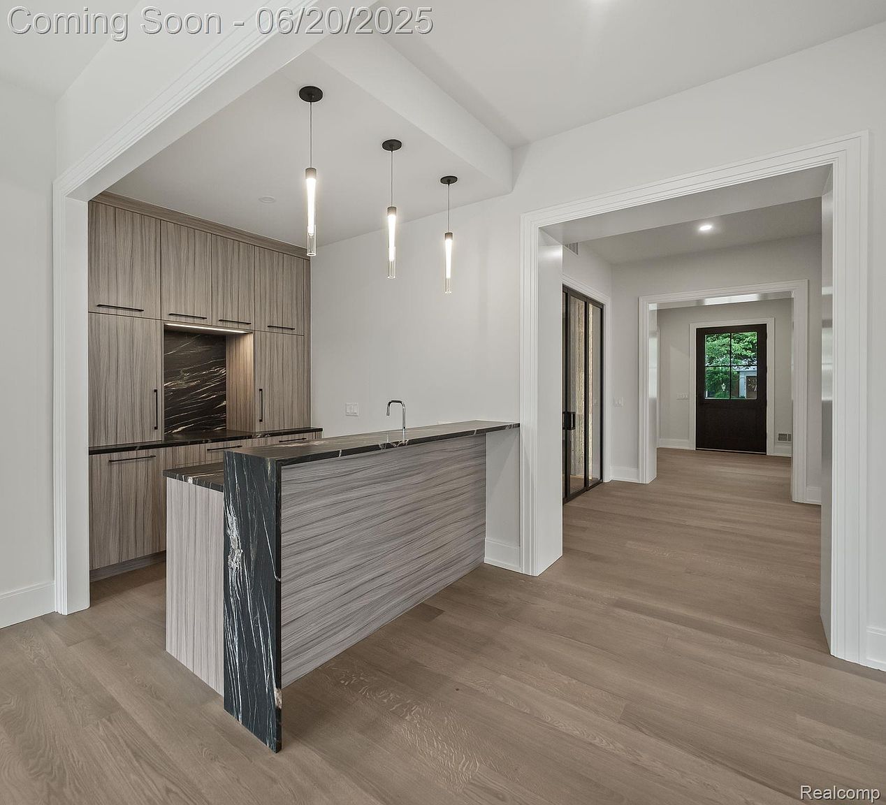 This interior shot showcases a modern kitchen with sleek, light wood cabinetry and a striking island featuring a black marble accent. Pendant lights hang above the island, adding a touch of elegance. The open layout flows seamlessly into a hallway, creating a spacious and inviting atmosphere.
