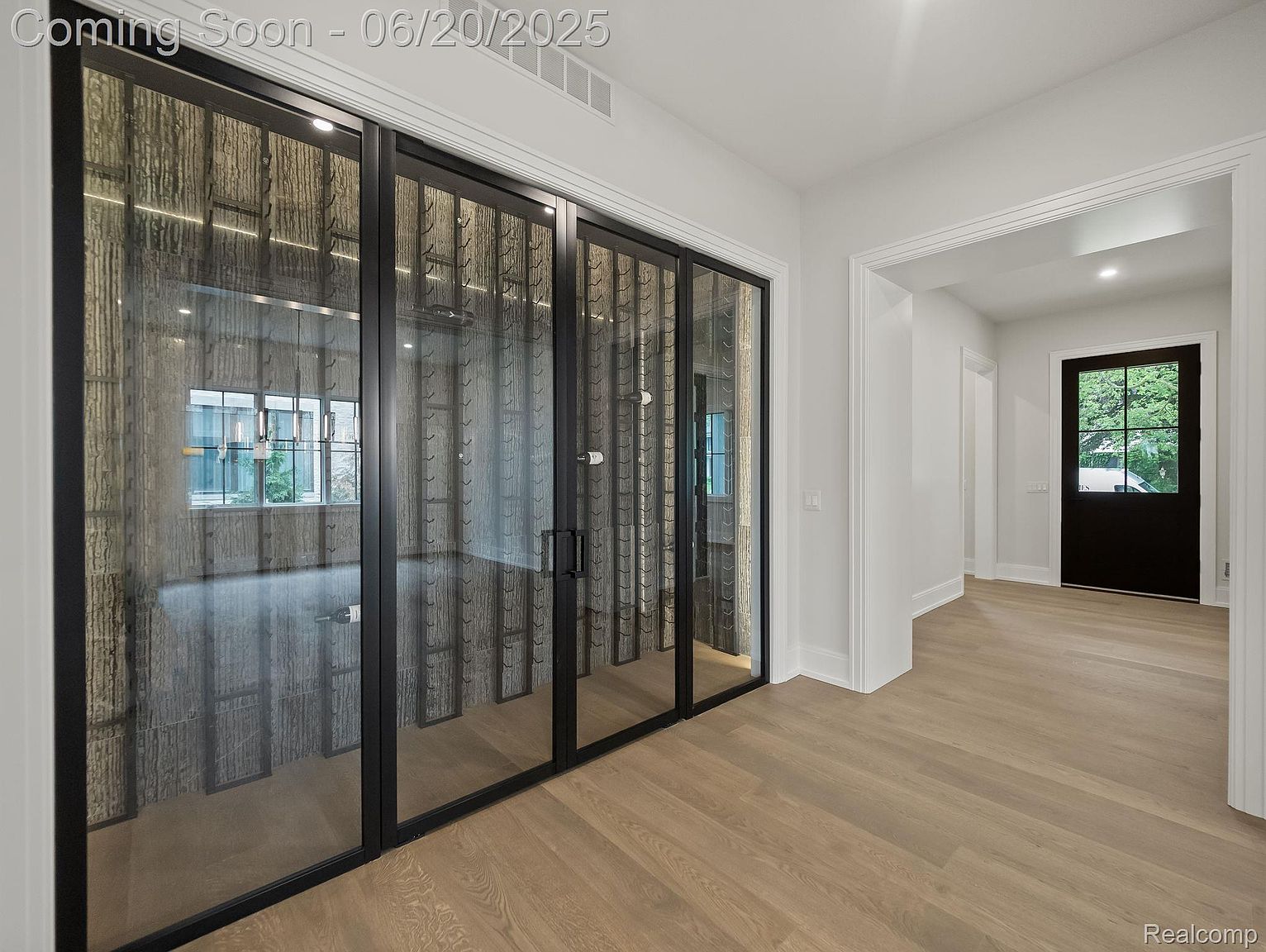 This interior shot showcases a modern hallway with light wood flooring and white walls. A striking wine cellar with black-framed glass doors is a prominent feature on the left. The hallway leads to a dark-colored door at the end, creating a sense of depth and inviting exploration.