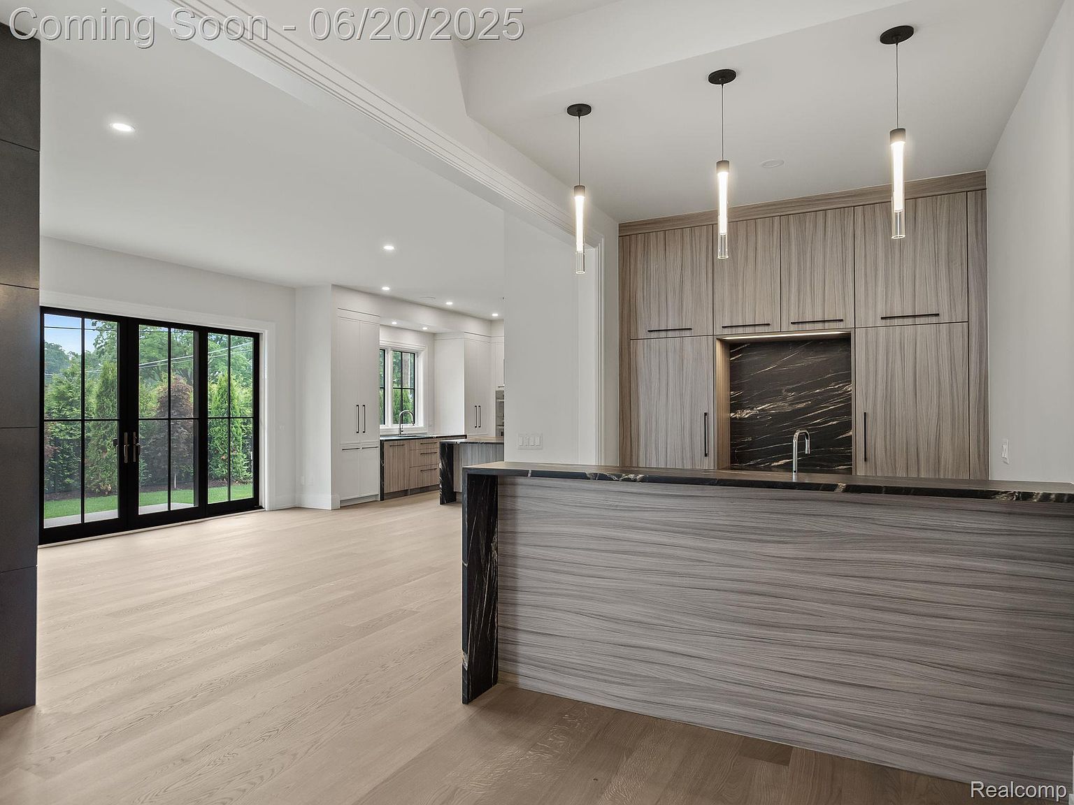 This interior shot showcases a modern kitchen with sleek, light wood cabinetry and a contrasting dark marble backsplash and countertop. The open floor plan leads to a bright living area with large glass doors, creating a seamless transition between indoor and outdoor spaces. The overall design exudes contemporary elegance and functionality.