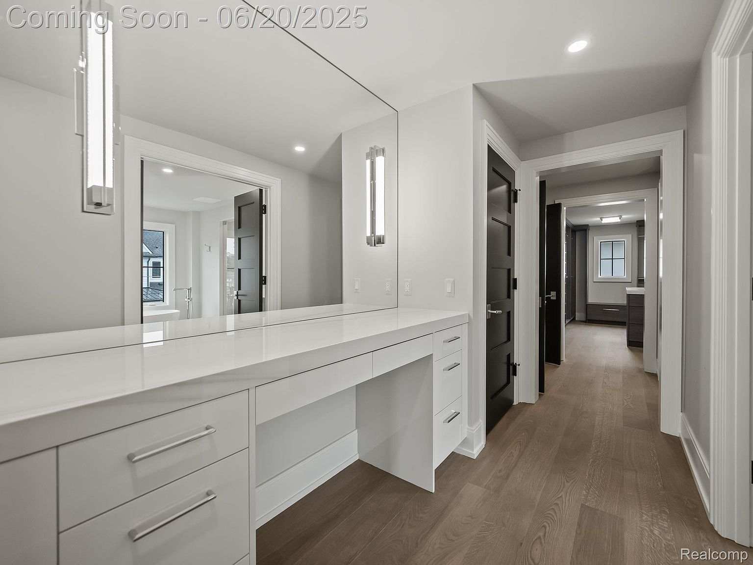 This is a well-lit primary bathroom featuring a long, white vanity with ample storage and a large mirror. The space has a modern aesthetic with clean lines and neutral tones, complemented by wood flooring. The view extends into a hallway with additional rooms, suggesting a spacious layout.