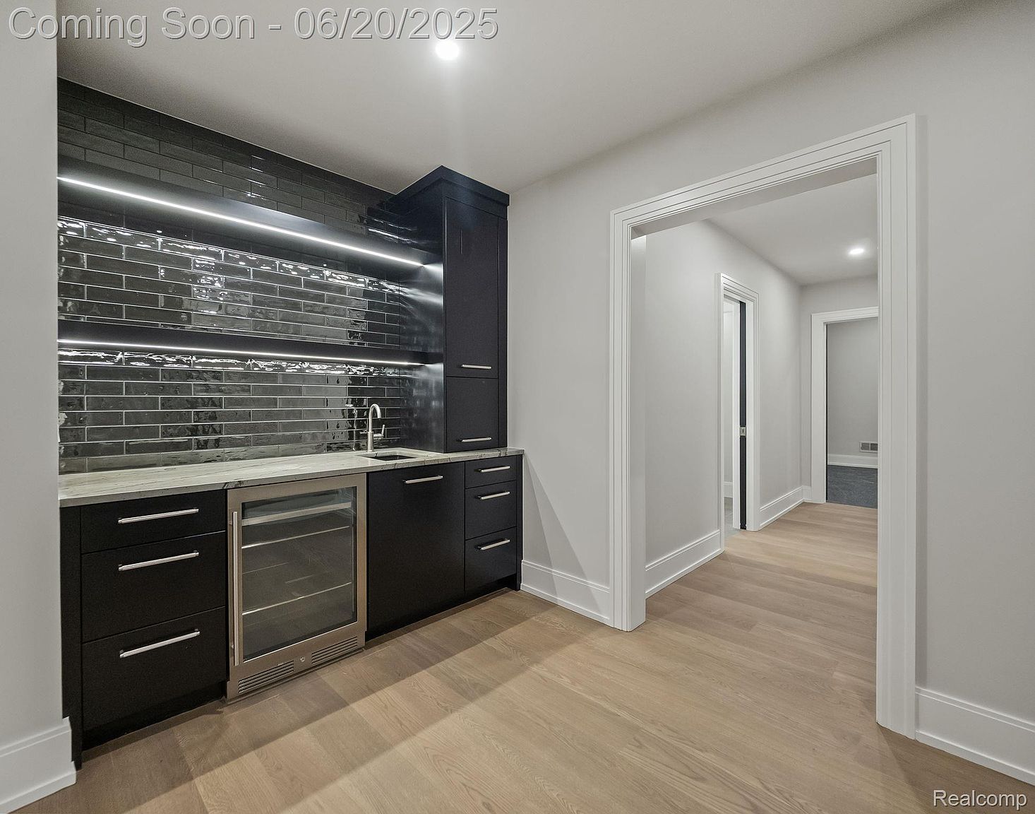 This interior shot showcases a modern wine cellar or bar area, featuring sleek black cabinetry, a built-in wine refrigerator, and a stylish black tile backsplash with integrated lighting. The light wood flooring and neutral gray walls create a sophisticated and inviting space, while the open doorway suggests a seamless transition to other areas of the home.