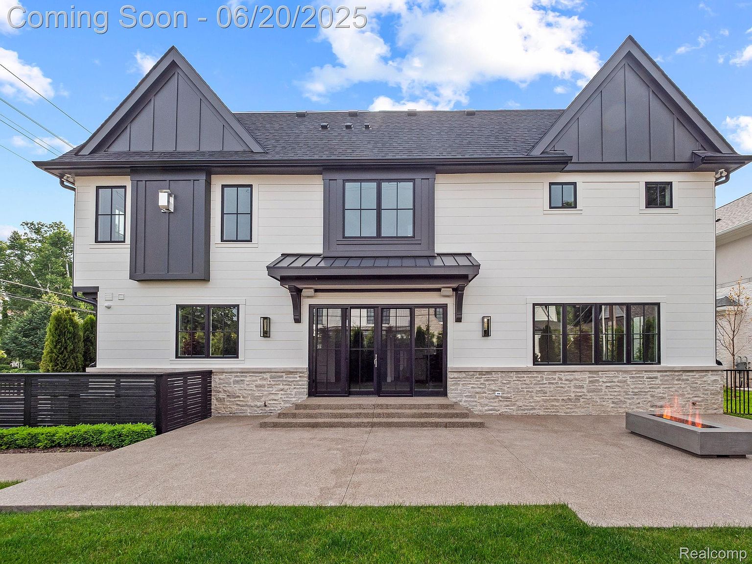 This is a front exterior view of a modern two-story house with a symmetrical design. The house features a white facade with black trim, including black-framed windows and dark gray gabled roof accents. A stone base adds texture, and a concrete patio with a fire pit extends the living space outdoors.
