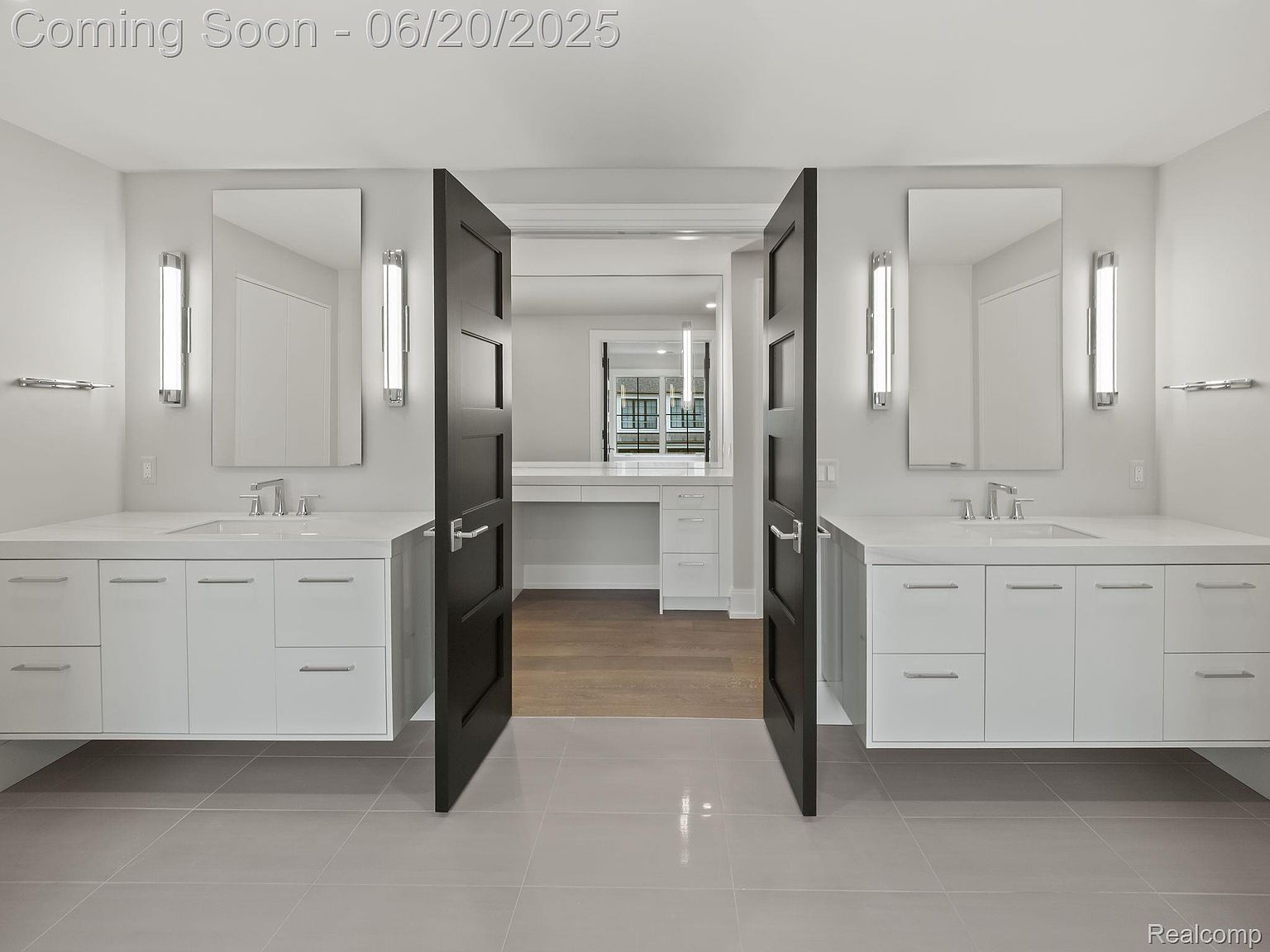 This is a modern primary bathroom featuring dual floating vanities with white countertops and cabinets, illuminated mirrors, and sleek fixtures. The bathroom has a contemporary design with gray tile flooring and dark-colored doors leading to another room, possibly a dressing area or closet. The overall impression is clean, spacious, and luxurious.