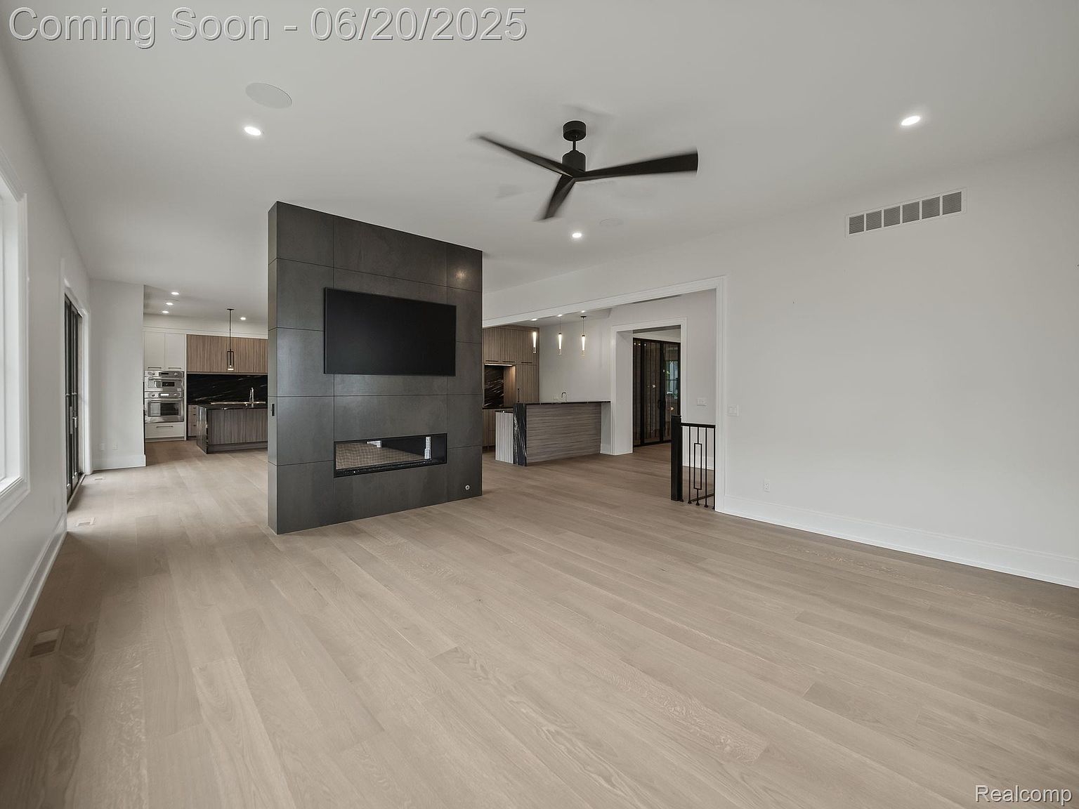 This is an interior shot of a modern living room featuring light wood flooring and a striking dark gray accent wall with a built-in fireplace and television. The open-concept design flows into the kitchen area, creating a spacious and inviting atmosphere. The room is well-lit with recessed lighting and a ceiling fan, enhancing the contemporary aesthetic.