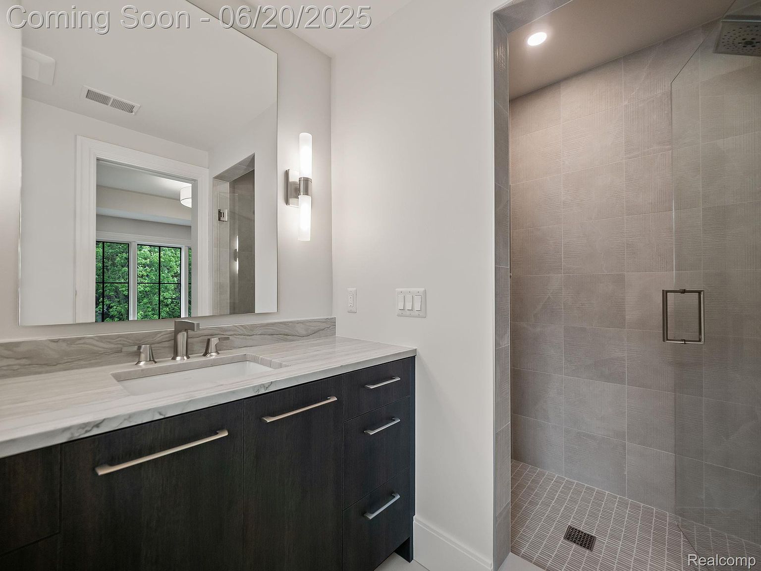 This is a modern bathroom featuring a dark wood vanity with a white marble countertop and brushed nickel hardware. A large mirror reflects the space, and a walk-in shower with gray tile and a glass door is visible to the right. The overall design is sleek and contemporary, creating a luxurious feel.