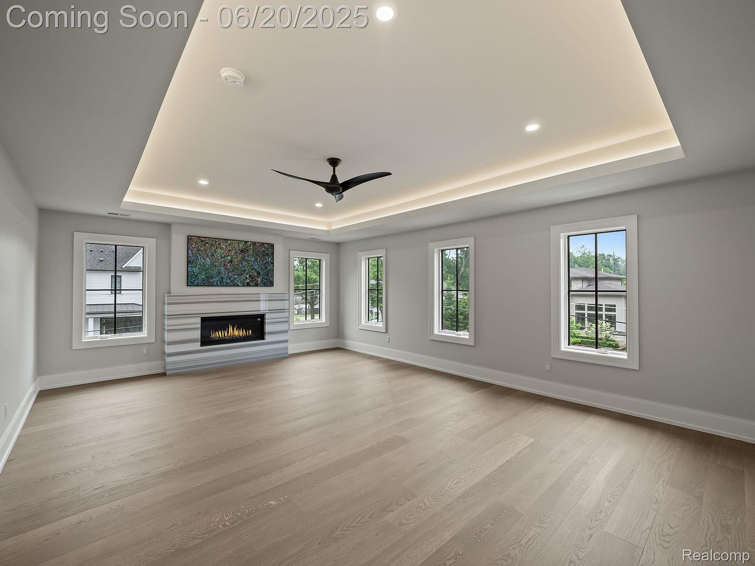 This is a spacious living room featuring light hardwood floors and neutral gray walls. A modern fireplace with a marble surround is the focal point, complemented by a large abstract artwork above. The room is well-lit with recessed lighting and natural light streaming through multiple windows, creating a bright and airy atmosphere.