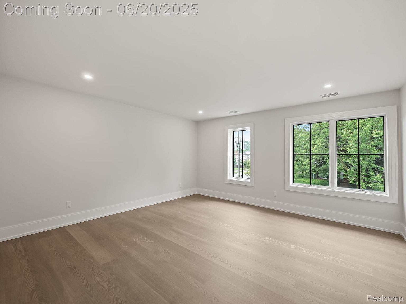 This is a bright and airy bedroom featuring light hardwood floors and neutral-colored walls. Two windows provide ample natural light and views of the outdoors. The room is spacious and appears to be newly constructed or renovated, offering a clean and modern aesthetic.