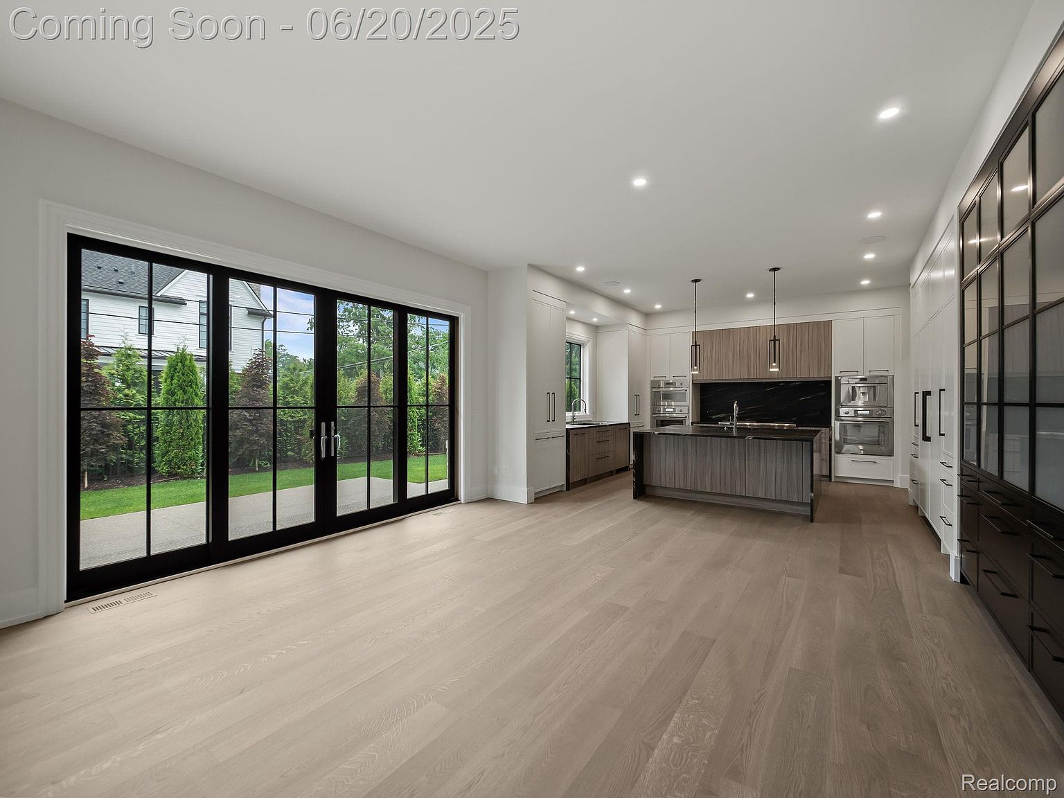 This is a bright and spacious open-concept kitchen and living area. The kitchen features a large island with dark countertops and light wood cabinetry, complemented by white cabinets and stainless steel appliances. Large black-framed sliding glass doors provide ample natural light and a view of the backyard, enhancing the modern and luxurious feel of the space.