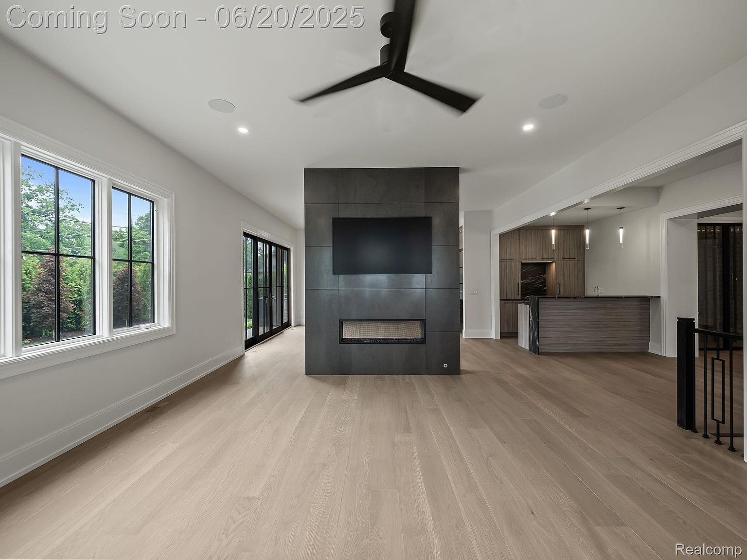 This is a spacious living room featuring light hardwood floors and a modern fireplace with a mounted television. Large windows provide ample natural light, and the open floor plan seamlessly connects to the kitchen area. The room exudes a contemporary and inviting atmosphere, perfect for relaxation and entertainment.