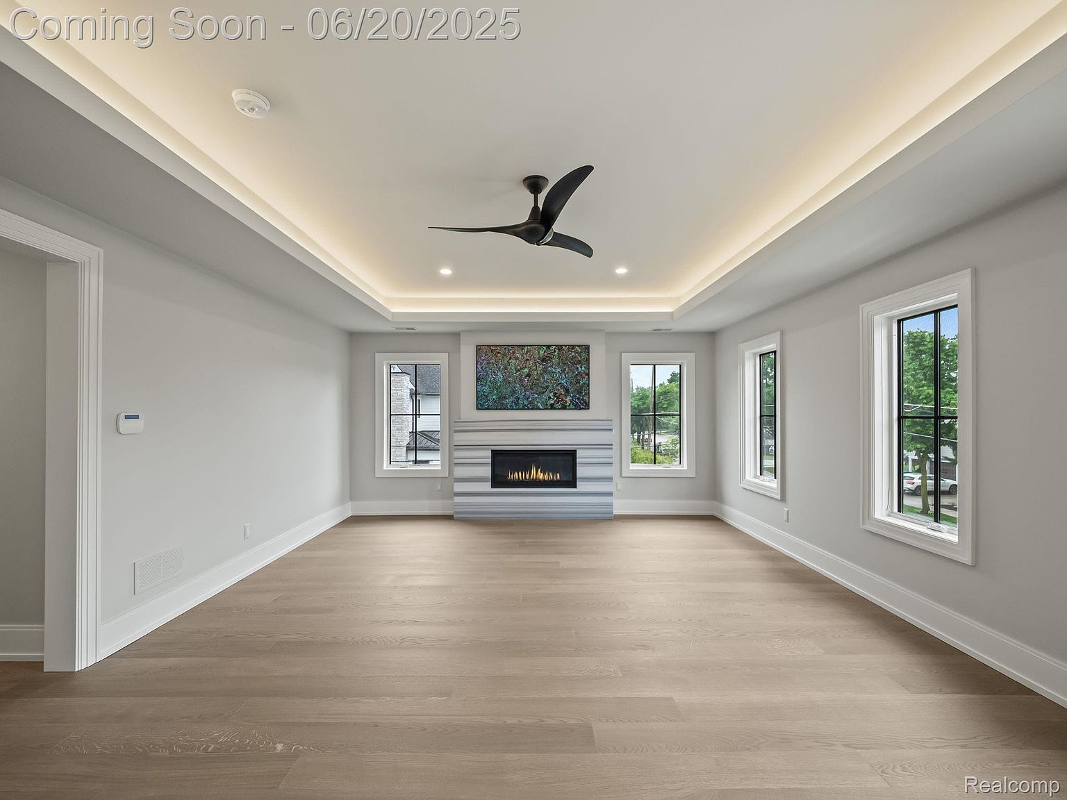 This is an interior shot of a living room featuring hardwood floors, a modern fireplace with a marble surround, and a ceiling fan. The room is well-lit with recessed lighting and natural light from the windows, creating a bright and airy atmosphere. The walls are painted in a neutral color, providing a blank canvas for potential buyers to envision their own decor.