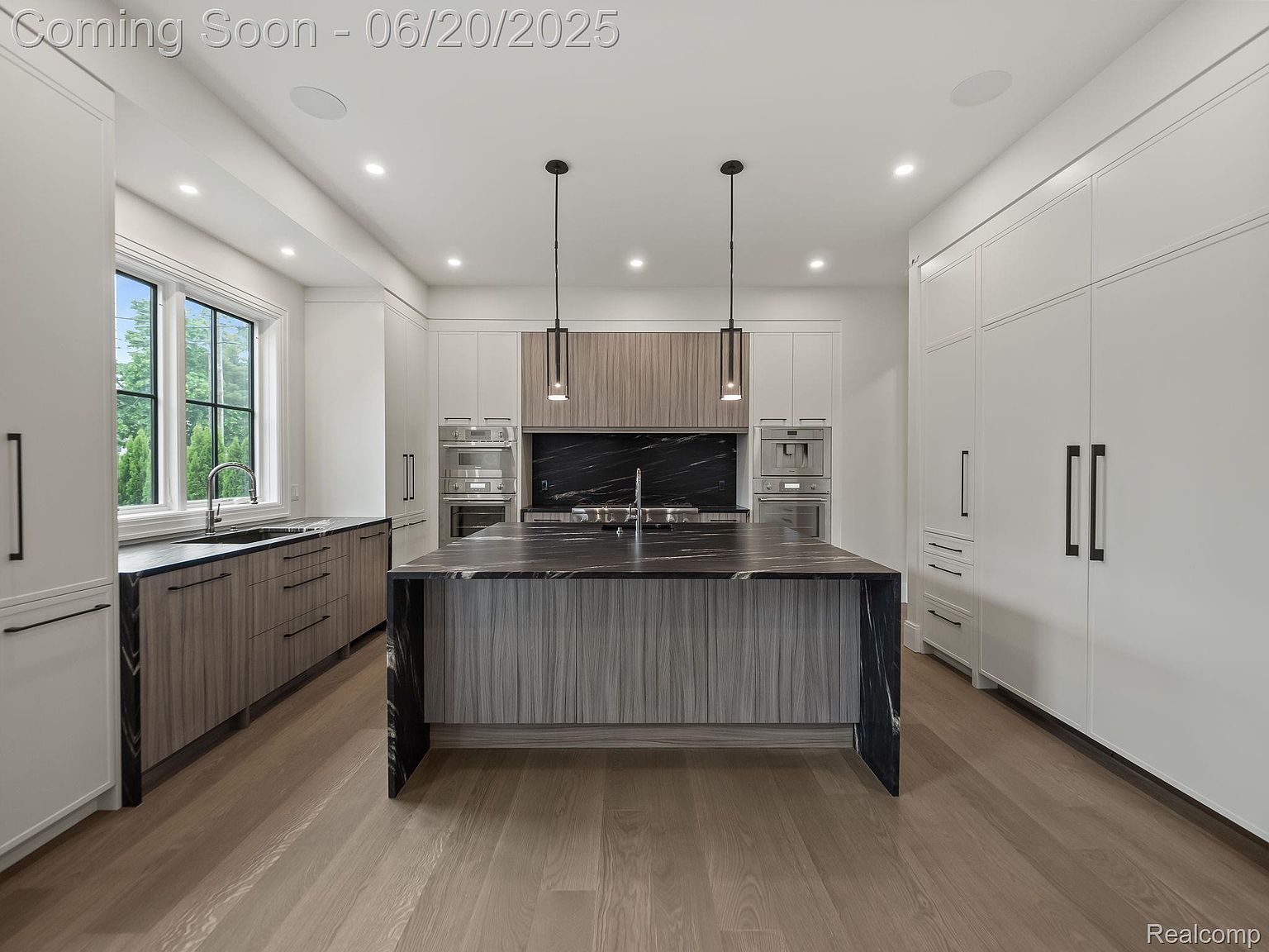 This is a bright and modern kitchen featuring a large island with a dark countertop and light wood-grain cabinetry. The kitchen also includes white cabinets, stainless steel appliances, and a black marble backsplash. The overall design is sleek and contemporary, creating a luxurious and functional space.