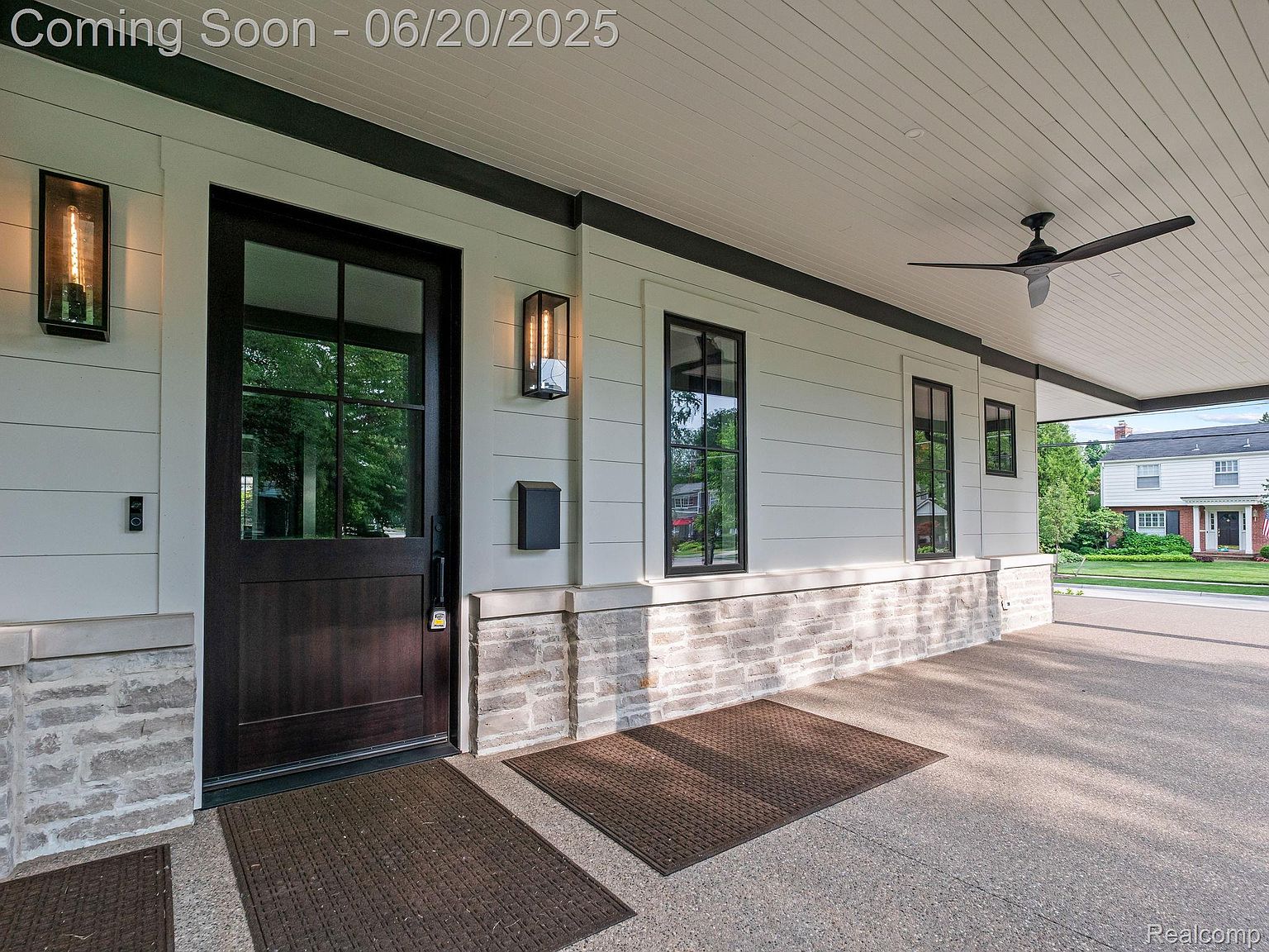 This is a welcoming entryway featuring a dark wood door with glass panes, flanked by modern light fixtures and a mailbox. The exterior is clad in white siding with a stone base, and a covered porch extends along the front. The scene is set during the day, with a view of the neighborhood across the street.