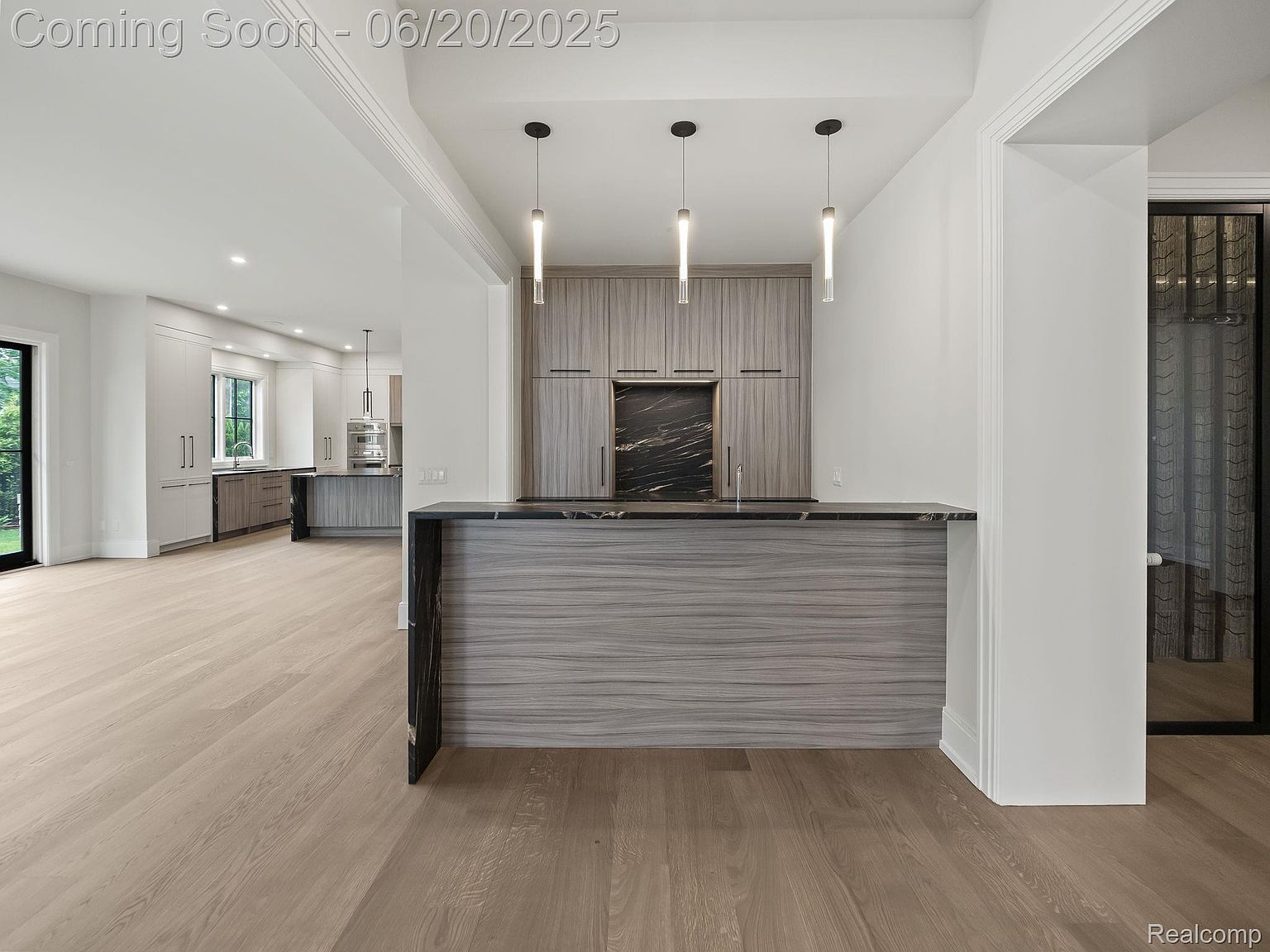 This interior shot showcases a modern kitchen with sleek, gray-toned cabinetry and black marble accents. Three pendant lights hang above a bar area, adding a touch of elegance. The open floor plan leads into a bright kitchen space with stainless steel appliances, creating a seamless and inviting atmosphere.