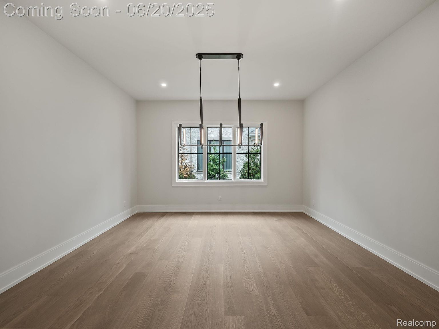 This is an interior shot of a dining room, showcasing a minimalist design with clean lines and neutral tones. The room features light wood flooring, white walls, and a modern chandelier hanging above where a dining table would be. A large window provides natural light and a view to the outside, enhancing the spacious feel of the room.
