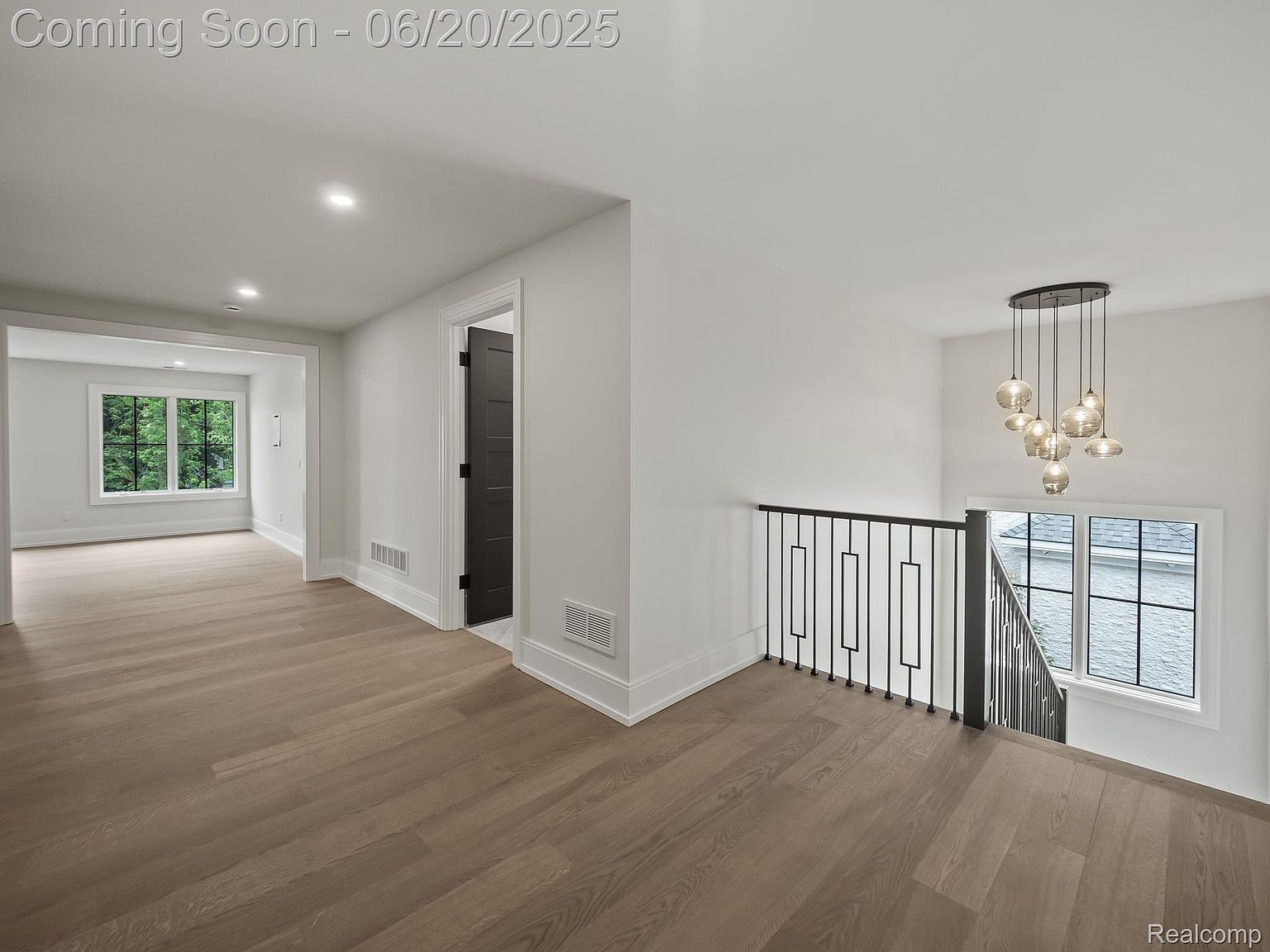 This interior shot showcases a bright and modern hallway with hardwood flooring and white walls. A black metal railing leads down the stairs, and a contemporary chandelier hangs above. The space is well-lit, creating a clean and inviting atmosphere.