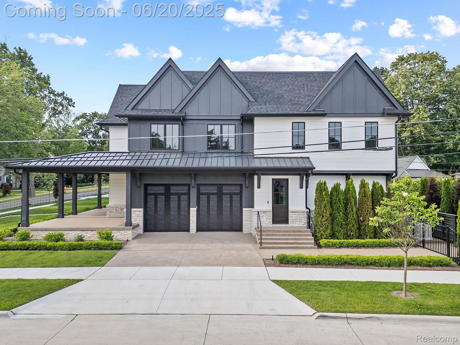 This is a front exterior view of a modern two-story home with a striking gray and white color scheme. The house features a covered porch on the left side, a two-car garage, and a well-manicured lawn with neatly trimmed hedges and a young tree. The architectural style is contemporary with gabled roof elements and black-framed windows, creating a sophisticated and inviting curb appeal.