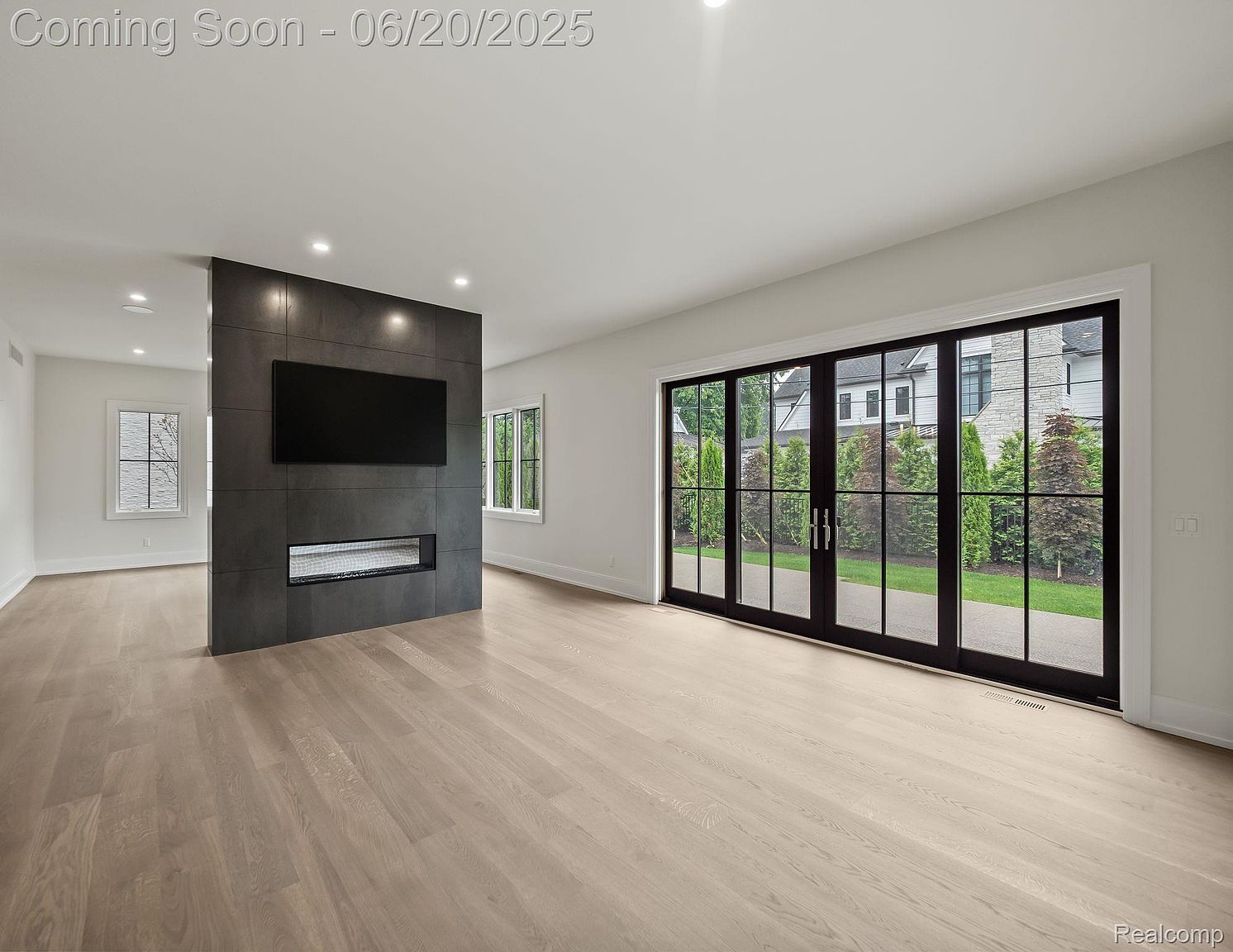 This is a bright and spacious living room featuring light hardwood floors and a modern fireplace with a mounted television. Large sliding glass doors provide ample natural light and a view of the exterior landscaping. The room's minimalist design and neutral color palette create a clean and inviting atmosphere.