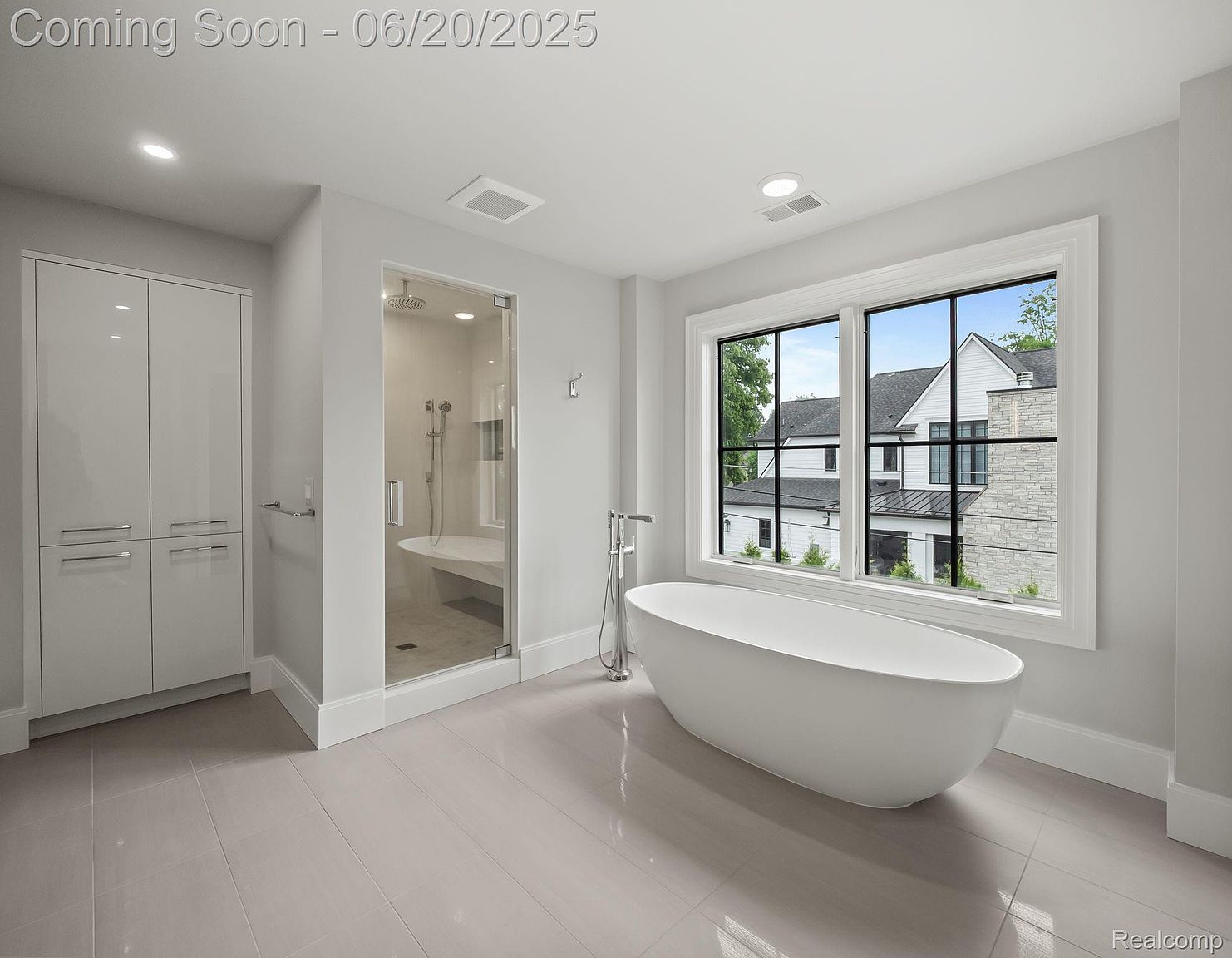 This is a bright and modern primary bathroom featuring a freestanding white bathtub positioned in front of a large window with black frames, offering an exterior view. The bathroom also includes a glass-enclosed shower with a built-in bench and a tall, sleek storage cabinet. The neutral color palette and clean lines create a spa-like atmosphere.