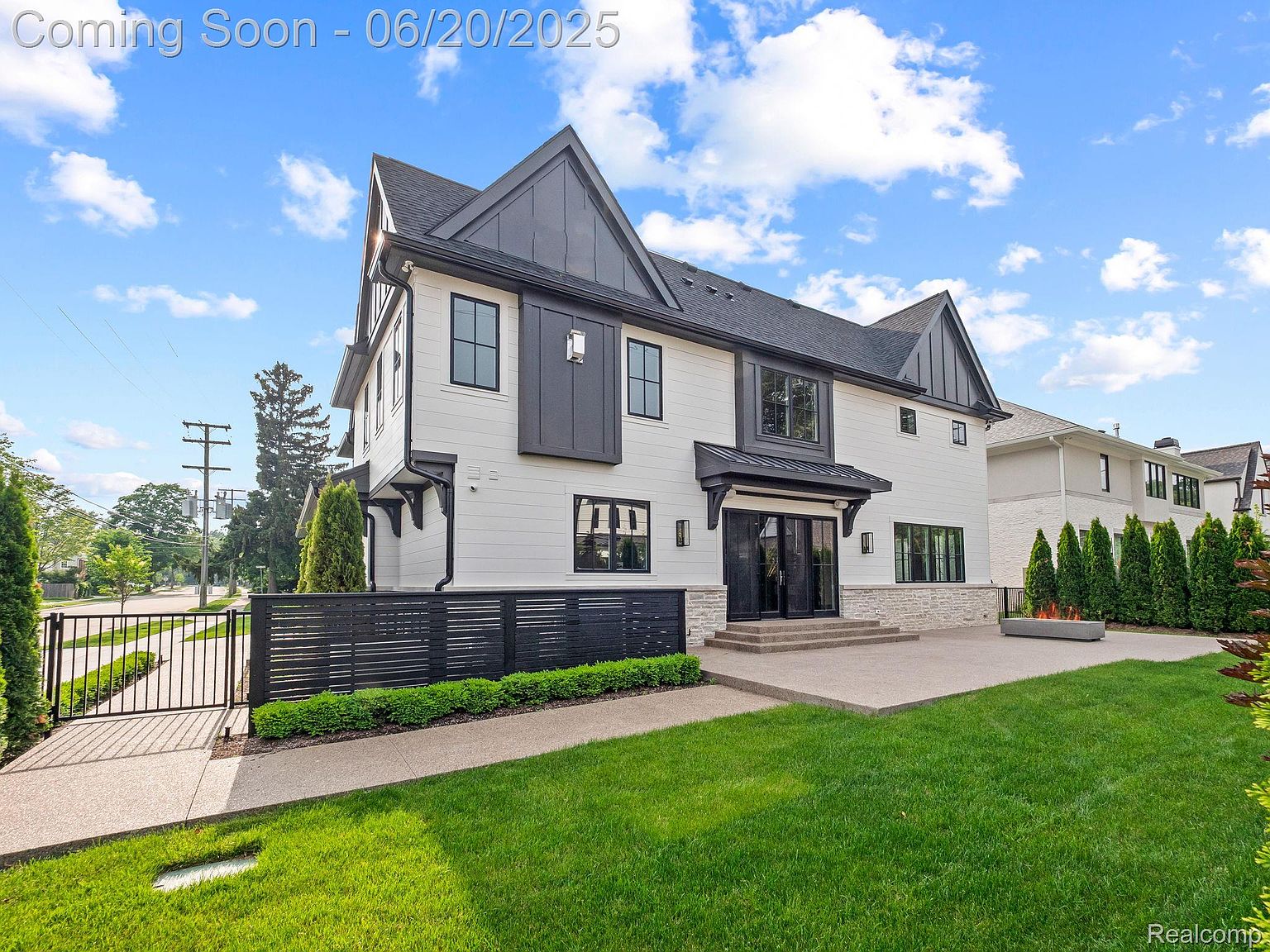 This is a front exterior view of a modern two-story house with a combination of white siding and dark gray accents. The house features a well-manicured lawn, a black horizontal fence, and a paved walkway leading to the entrance. The overall impression is one of contemporary elegance and curb appeal.