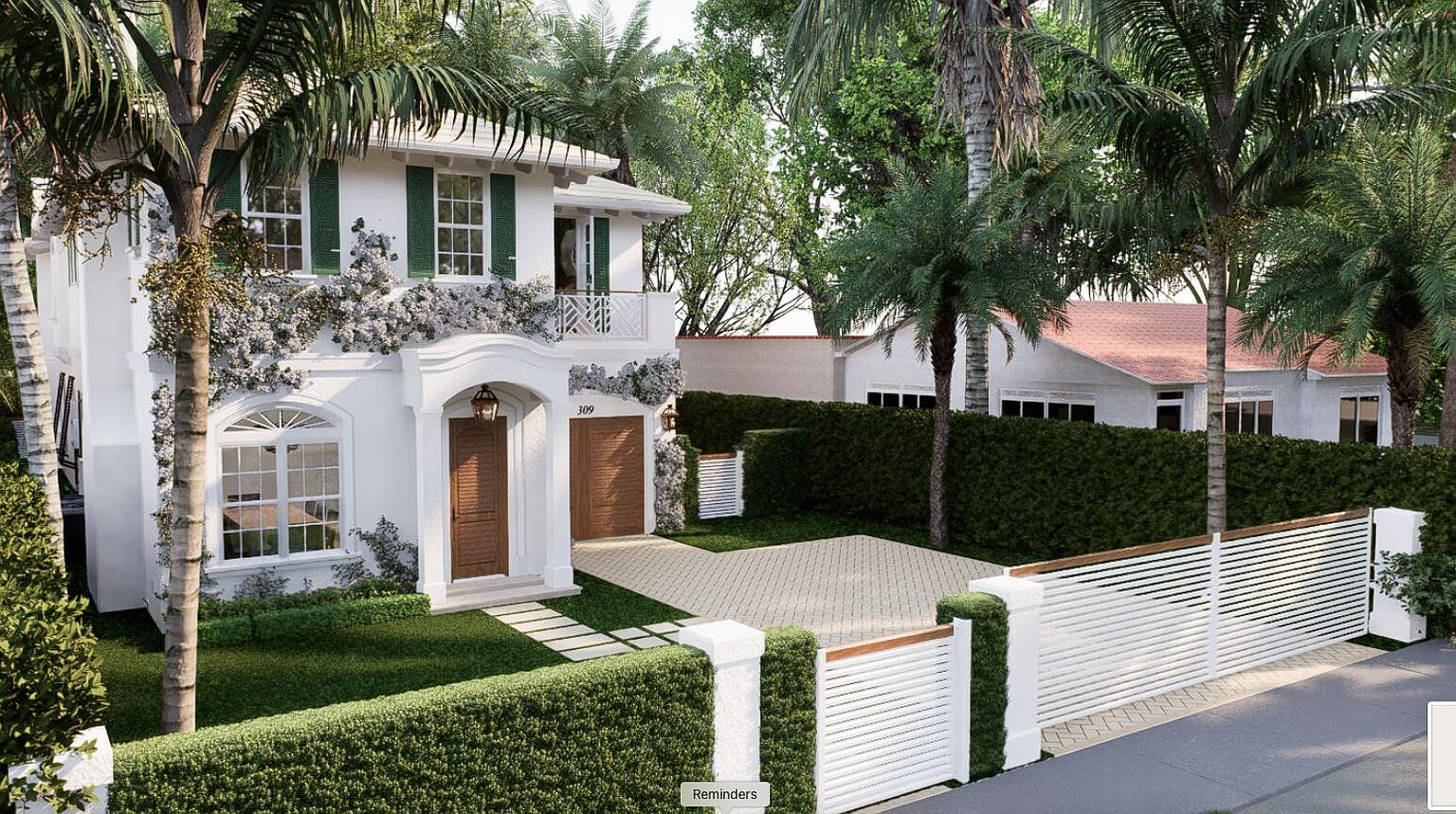 This is a front exterior view of a two-story white house with green shutters and a brown garage door. The house is surrounded by lush greenery, including palm trees and manicured hedges. A white fence with wooden accents encloses the property, creating a sense of privacy and elegance.