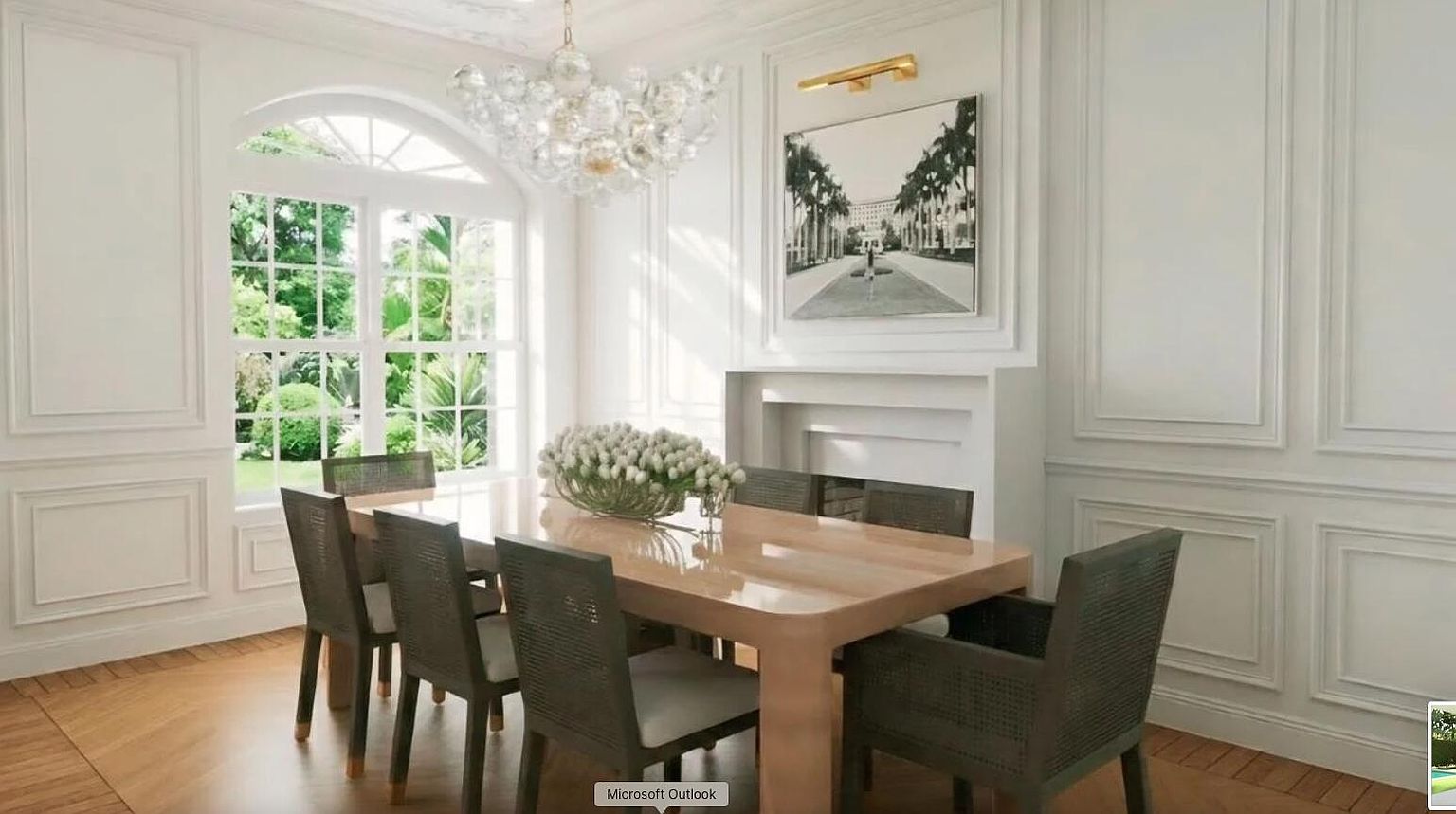 This is an elegant dining room featuring white paneled walls, a large window with a view of lush greenery, and a modern chandelier. A light wood dining table is surrounded by upholstered chairs, and a piece of art hangs above a fireplace. The room exudes a sophisticated and bright atmosphere.