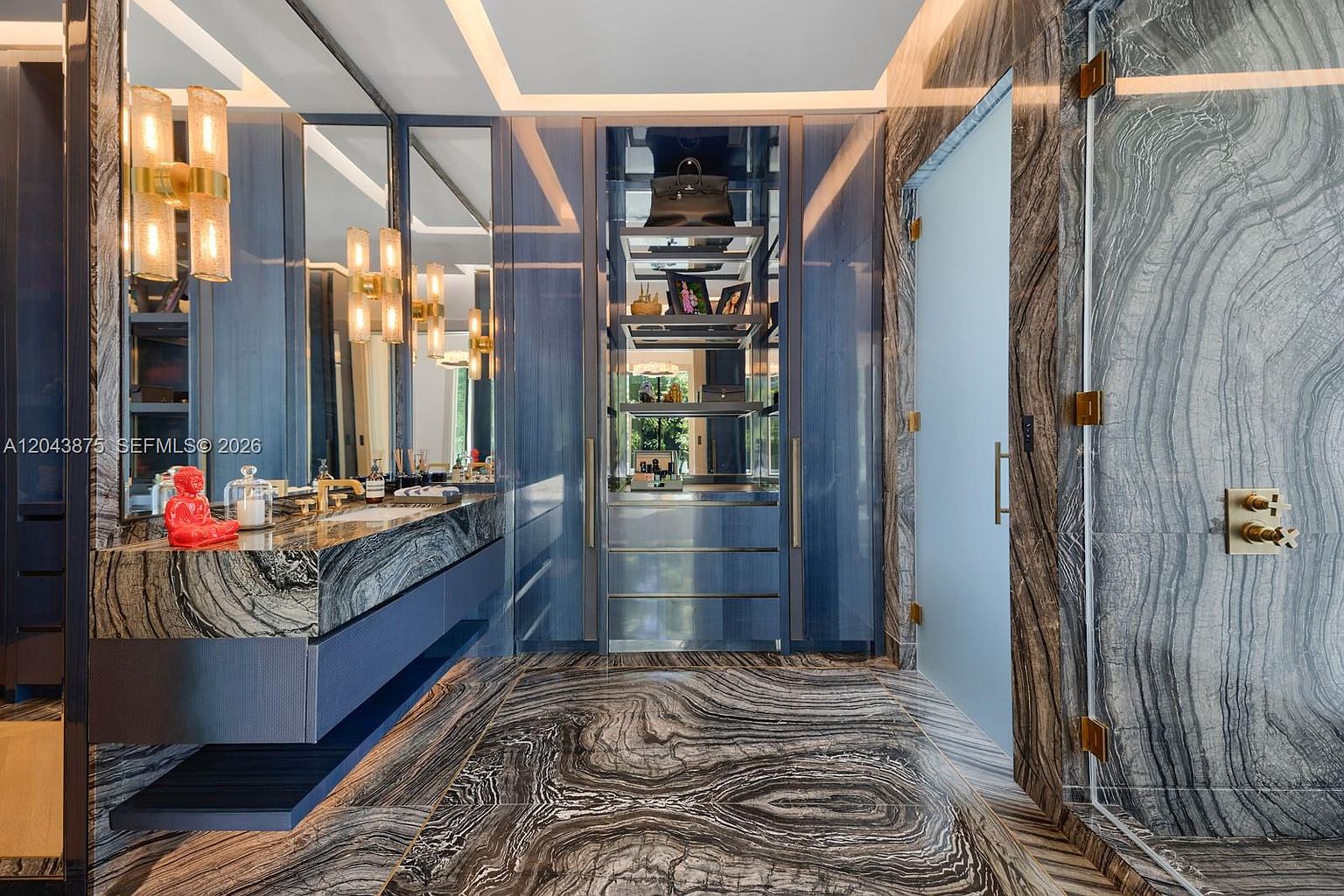 This luxurious primary bathroom features dramatic, dark-veined stone surfaces on the floors and walls, creating a sophisticated and moody aesthetic. A sleek vanity with a stone countertop is paired with blue cabinetry and modern gold-toned fixtures, while a central open shelving unit provides elegant storage. The space is illuminated by warm, vertical wall sconces and recessed lighting, reflecting off the polished surfaces to enhance the high-end, contemporary design.
