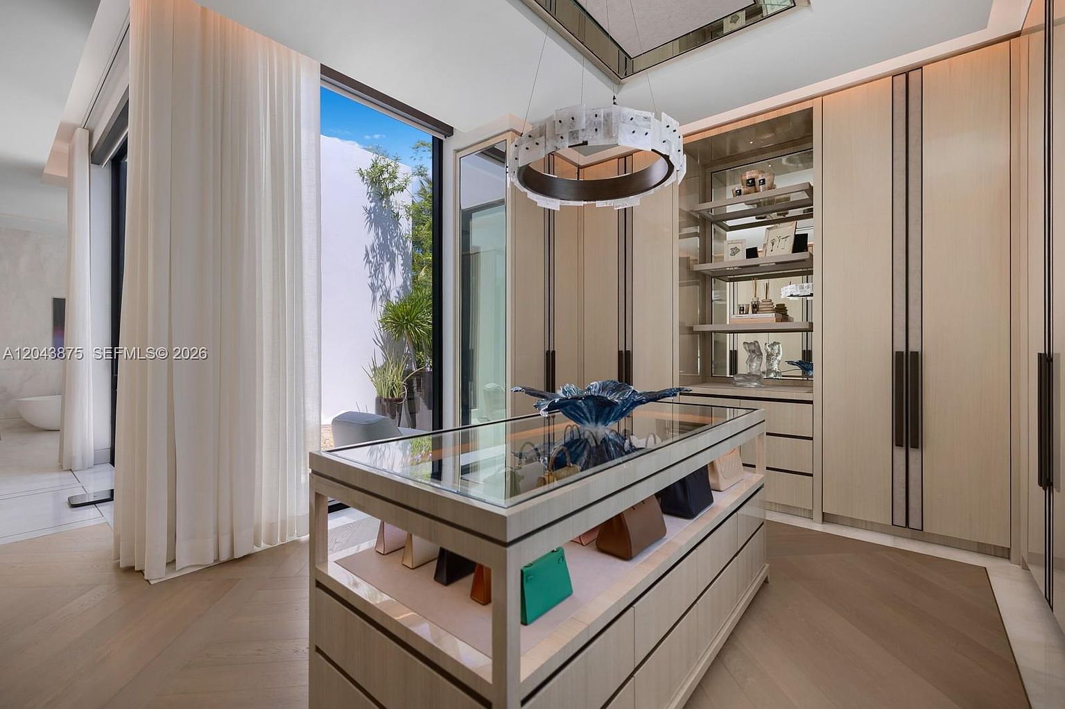 This luxurious walk-in closet features custom light wood cabinetry with integrated shelving and a central glass-topped island display for accessories. A modern circular chandelier hangs above the island, while large glass doors provide a seamless transition to an outdoor garden area. The space is finished with light-toned chevron-patterned wood flooring, creating an elegant and organized boutique-like atmosphere.