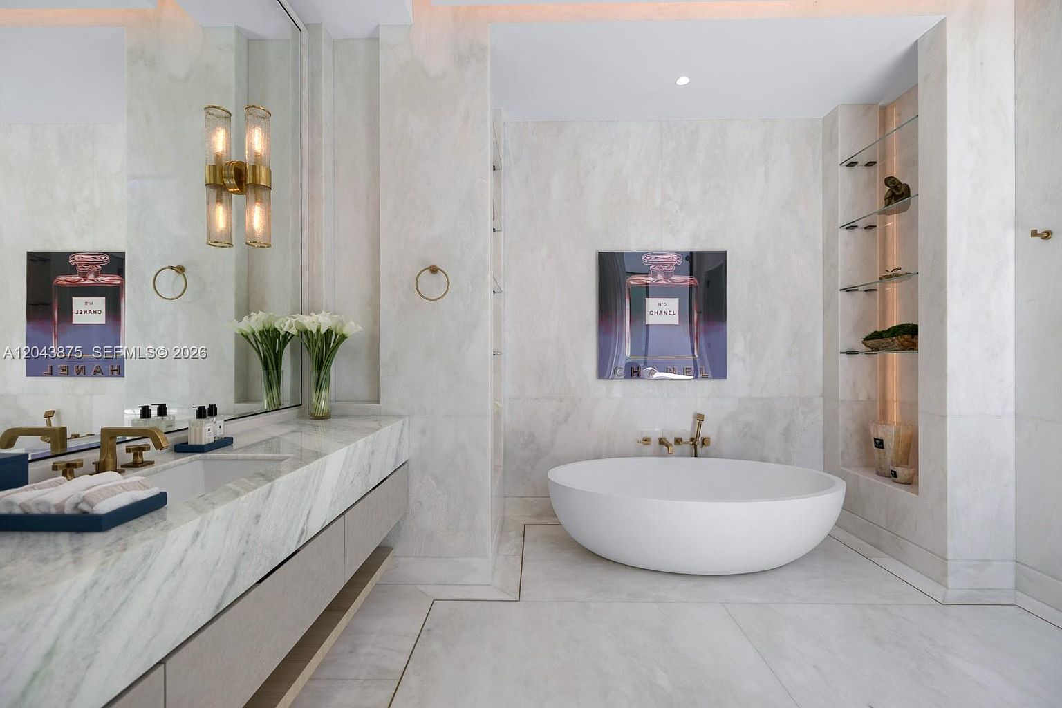 This luxurious primary bathroom features a sleek, white freestanding soaking tub centered against a light-toned stone wall adorned with a large Chanel No. 5 artwork. To the left, a long vanity with a stone countertop and gold-toned fixtures is complemented by a large mirror and elegant wall sconces. The space is finished with integrated shelving and light-colored stone flooring, creating a sophisticated and high-end spa-like atmosphere.