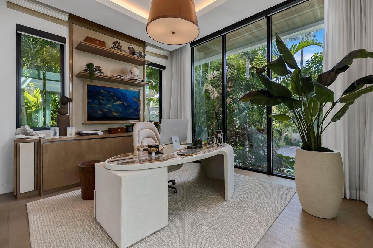 This sophisticated home office features a unique, curved desk with a patterned stone top positioned centrally in the room. Large windows provide an expansive view of lush, tropical greenery outside, creating a serene and bright atmosphere. The space is accented by built-in shelving, a modern pendant light, and a large potted plant, all contributing to a refined and professional aesthetic.