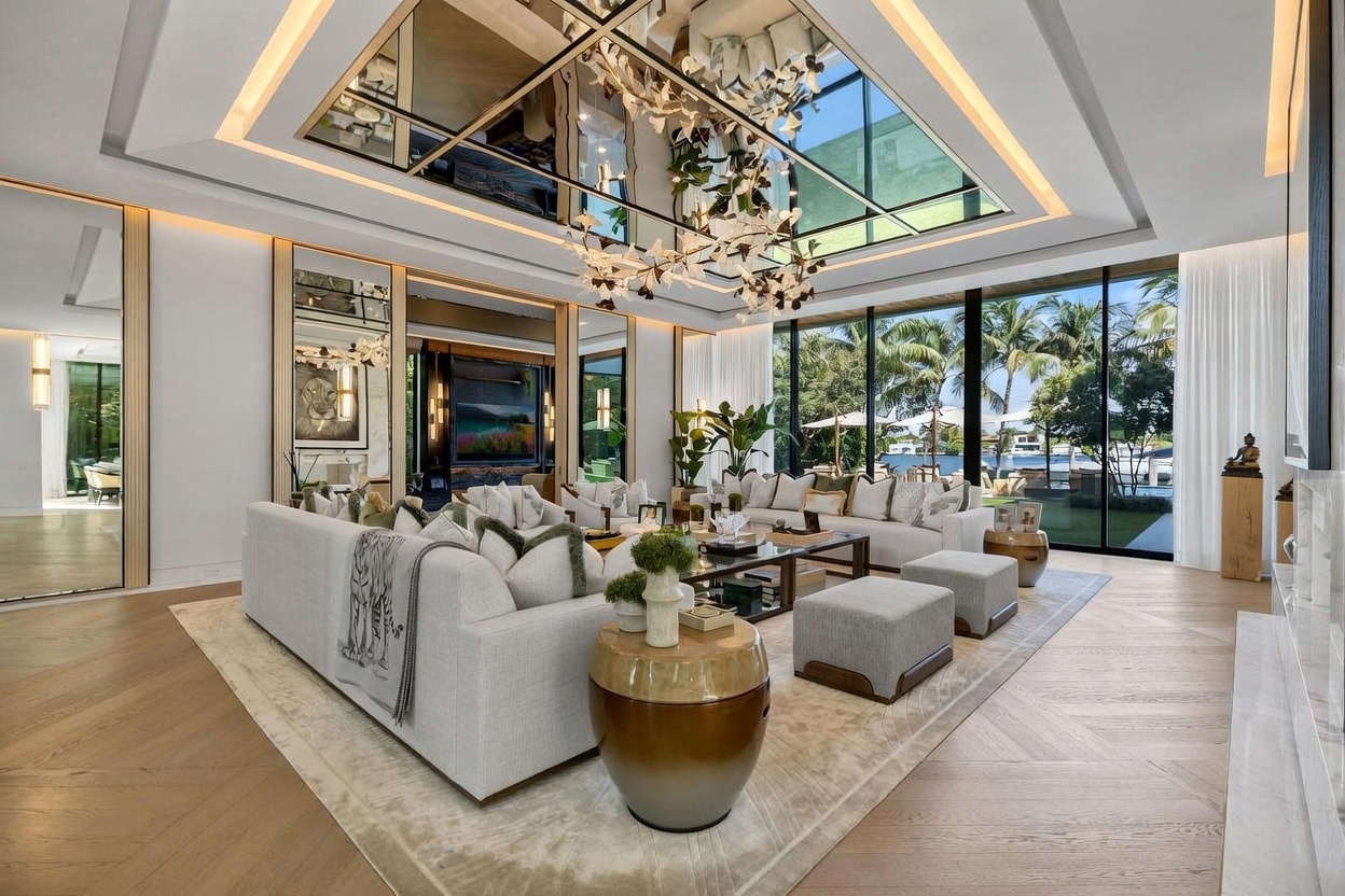 This expansive, high-end living room features a sophisticated open-concept design with light wood flooring arranged in a chevron pattern. The space is anchored by a large, plush sectional sofa and a central coffee table, all positioned beneath a striking, modern chandelier and a mirrored ceiling feature. Large windows provide an unobstructed view of the lush outdoor landscape and waterfront, creating a seamless transition between indoor luxury and the natural environment.