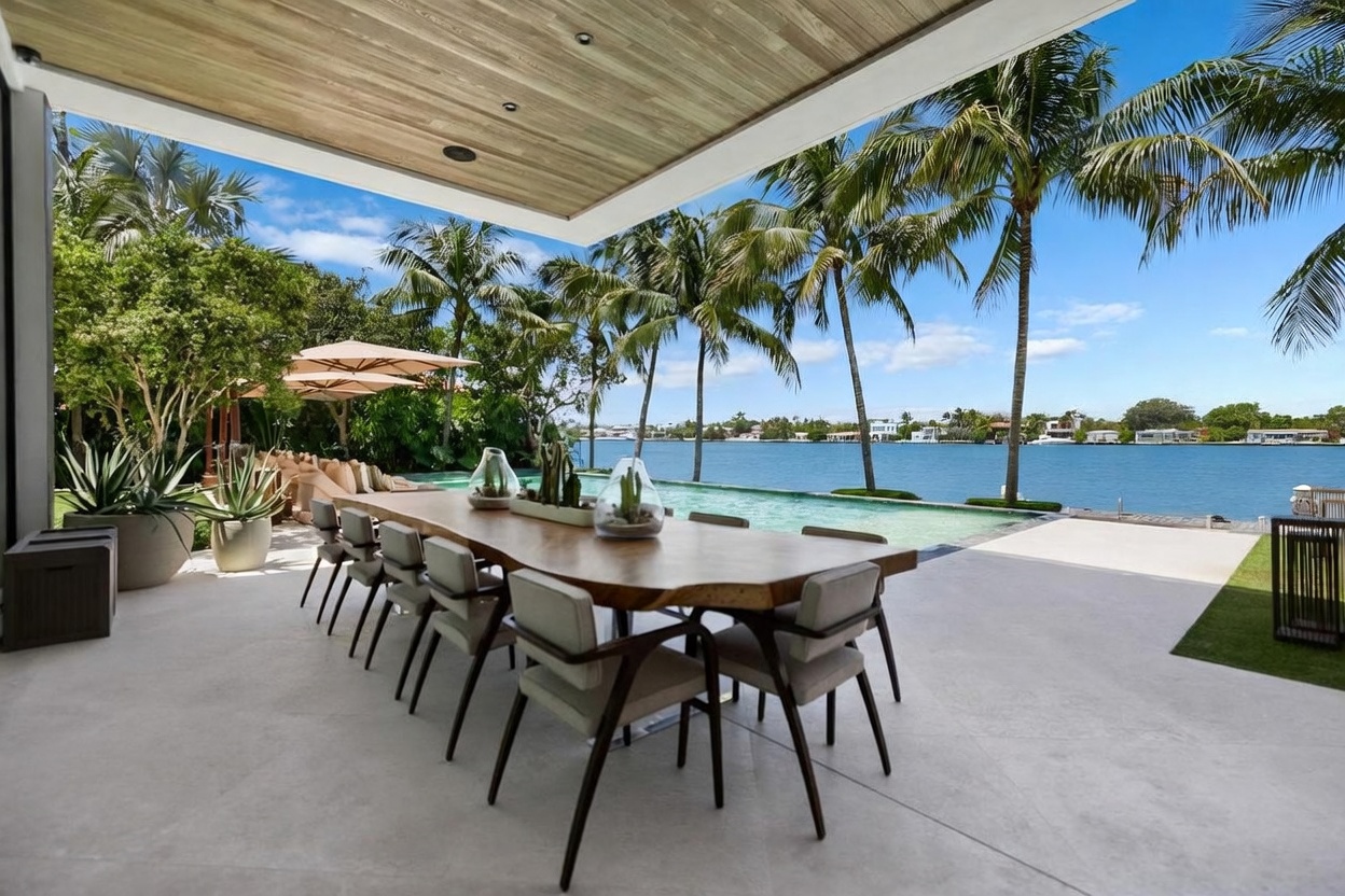 This expansive covered outdoor dining area features a large, natural-edge wood table surrounded by modern, neutral-toned chairs. The space seamlessly transitions to an infinity-edge pool that overlooks a wide waterway, framed by tall palm trees and a clear blue sky. The architectural design emphasizes an open-air connection between the shaded patio and the waterfront landscape, creating a luxurious and serene atmosphere.