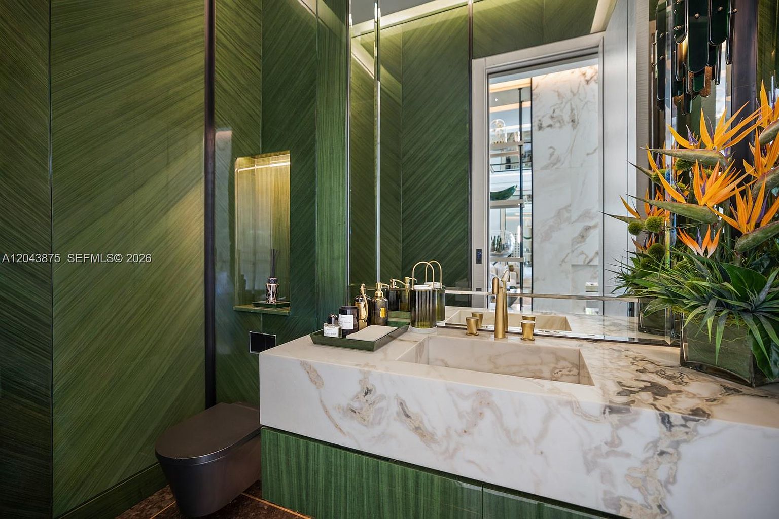 This sophisticated powder room features striking green wood-paneled walls with a diagonal grain pattern and a luxurious stone vanity top with an integrated sink. A large mirror reflects the space, while gold-toned fixtures and a vibrant floral arrangement add a touch of elegance. The room is completed by a dark-toned toilet and recessed lighting, creating a moody and high-end atmosphere.