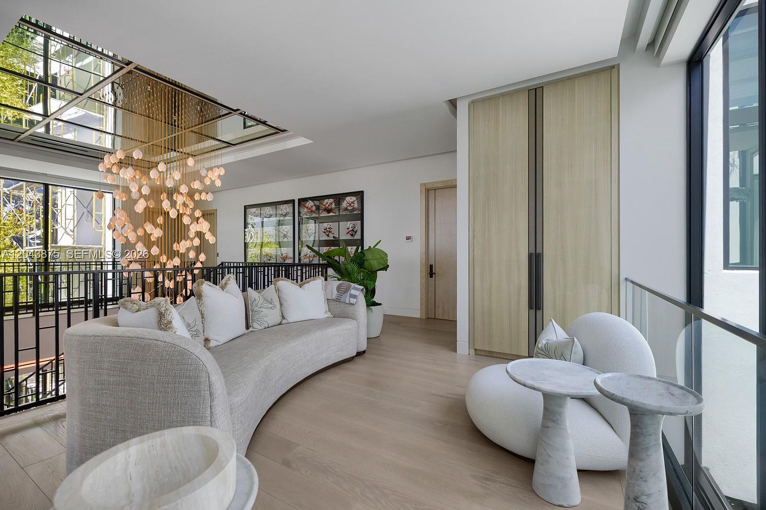 This elegant, light-filled living area features a curved sofa with plush pillows and a modern, rounded accent chair paired with two small stone-topped tables. A dramatic, multi-tiered chandelier hangs from the ceiling, casting a warm glow over the space, while a glass railing overlooks the level below. The room is finished with light wood flooring and neutral walls, creating a sophisticated and airy atmosphere.