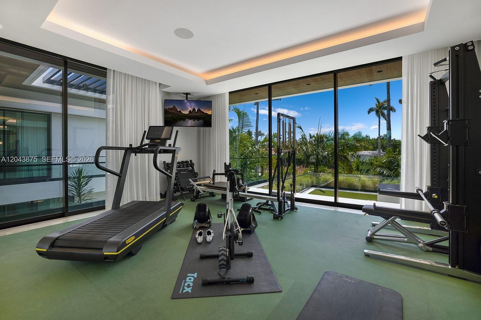 This modern home gym features a sleek, minimalist design with green flooring and expansive floor-to-ceiling glass walls that provide an unobstructed view of the lush outdoor landscape. The room is equipped with professional-grade fitness equipment, including a treadmill, a stationary bike on a branded mat, and a multi-functional weight station. The space is illuminated by recessed lighting and a contemporary tray ceiling, creating a bright and motivating environment for exercise.