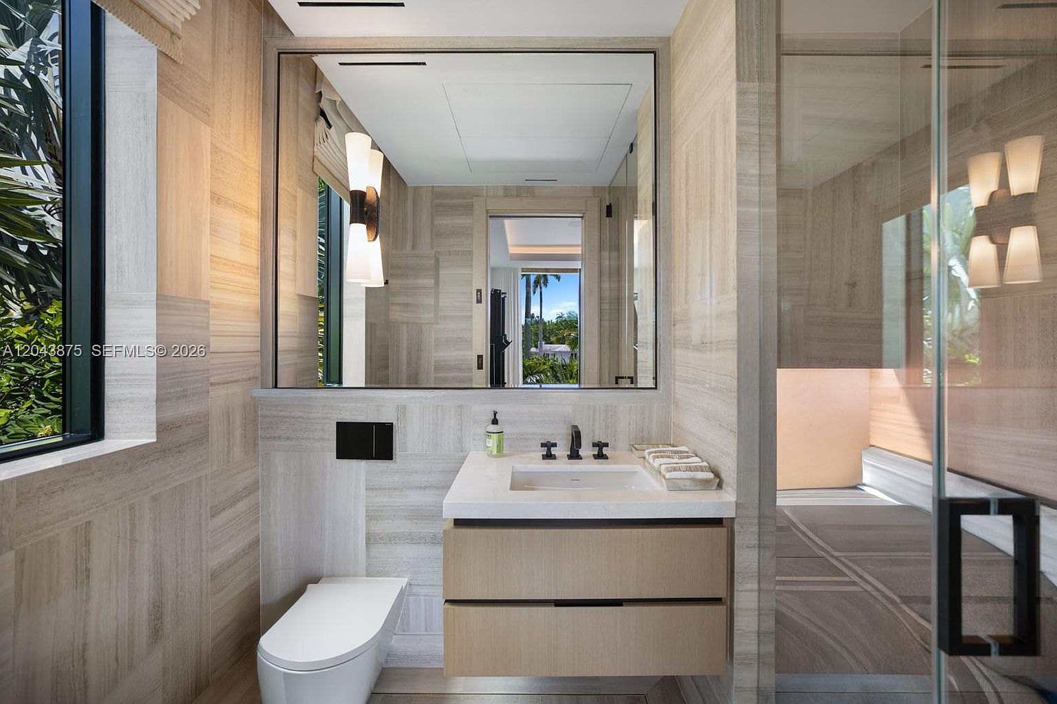 This modern bathroom features a floating vanity with light wood cabinetry and a stone countertop, complemented by a wall-mounted toilet and a large, sleek mirror. The walls are clad in neutral, linear-patterned stone tiles that create a cohesive and sophisticated aesthetic. A tall window to the left provides natural light, while a glass-enclosed shower area is visible to the right, emphasizing a clean and minimalist design.