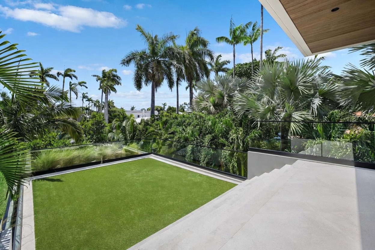 This elevated terrace features a sleek, modern design with a glass railing that provides an unobstructed view of the lush, tropical landscaping below. A vibrant green artificial turf lawn occupies the center of the deck, bordered by a stone-tiled patio area and a wooden ceiling overhang. The perspective captures a serene, high-end outdoor living space surrounded by mature palm trees under a bright blue sky.