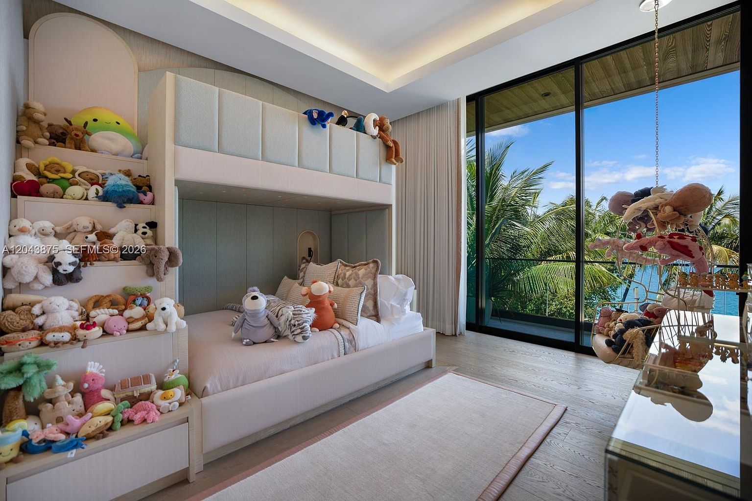 This bright and playful child's bedroom features a custom-built bunk bed unit with integrated shelving stairs filled with plush toys. The room is anchored by a light-toned area rug on wood flooring and includes a hanging chair near the large glass doors that provide a scenic view of palm trees and the water beyond. The space is designed with a soft, neutral color palette and clean lines, creating a serene yet whimsical atmosphere.