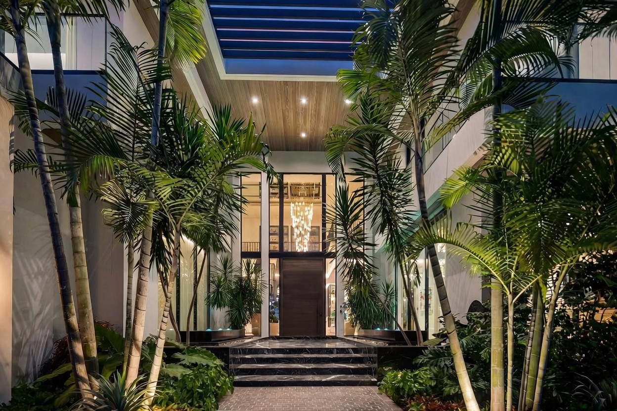 This grand entryway features a dramatic, symmetrical design with a central wooden door framed by expansive glass panels that reach the ceiling. The entrance is elevated by a set of wide, dark stone steps and flanked by lush palm trees and manicured landscaping, creating a luxurious, resort-like atmosphere. Overhead, a slatted architectural ceiling detail with recessed lighting adds depth and a modern aesthetic to the covered approach.